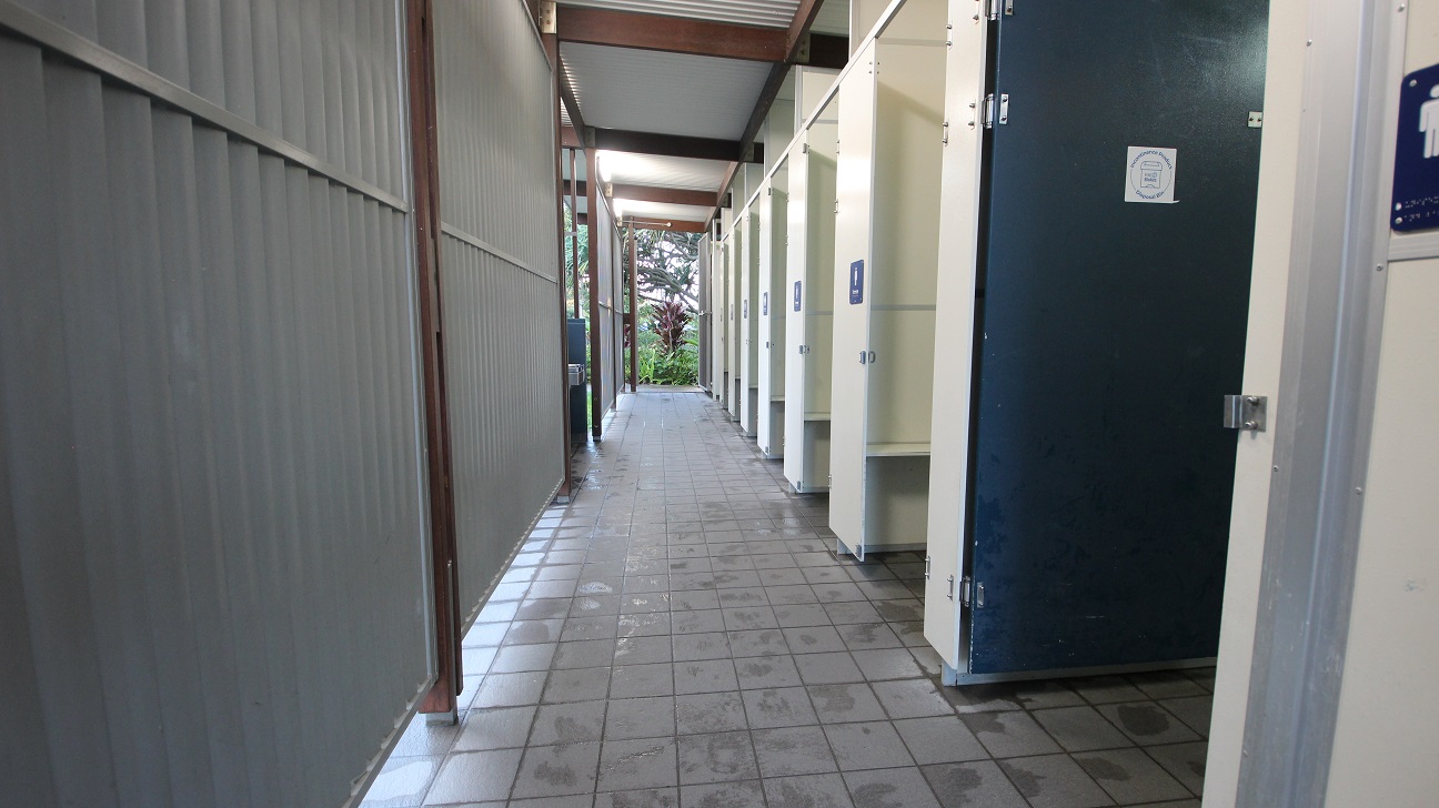 A corridor with a tiled floor and a corrugated metal ceiling. On the right, there are several toilet cubicles with white doors, one of which is dark blue and slightly ajar. Each door has a sign indicating a toilet. On the left, a grey corrugated wall runs the length of the corridor. At the end of the corridor, there is an opening leading to an outdoor area with visible greenery. A corridor with a tiled floor and a corrugated metal ceiling. On the right, there are several toilet cubicles with white doors, one of which is dark blue and slightly ajar. Each door has a sign indicating a toilet. On the left, a grey corrugated wall runs the length of the corridor. At the end of the corridor, there is an opening leading to an outdoor area with visible greenery.