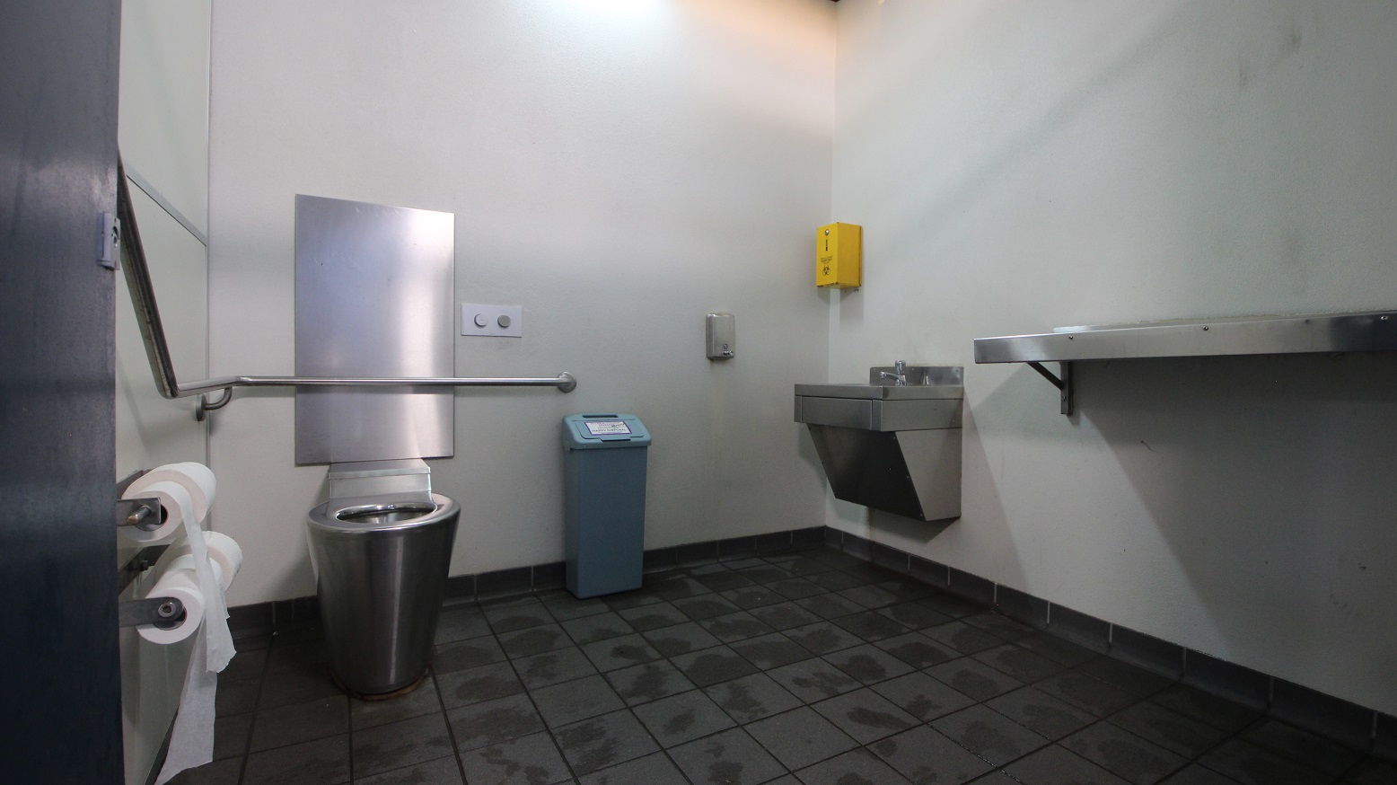A public restroom with a stainless steel toilet on the left, equipped with a grab rail and toilet paper holder. A blue bin is positioned nearby. On the right, a stainless steel sink is mounted on the wall, with a yellow dispenser above it. A metal shelf is also attached to the right wall. The floor is covered with dark tiles, and the walls are plain and light-coloured. A public restroom with a stainless steel toilet on the left, equipped with a grab rail and toilet paper holder. A blue bin is positioned nearby. On the right, a stainless steel sink is mounted on the wall, with a yellow dispenser above it. A metal shelf is also attached to the right wall. The floor is covered with dark tiles, and the walls are plain and light-coloured.