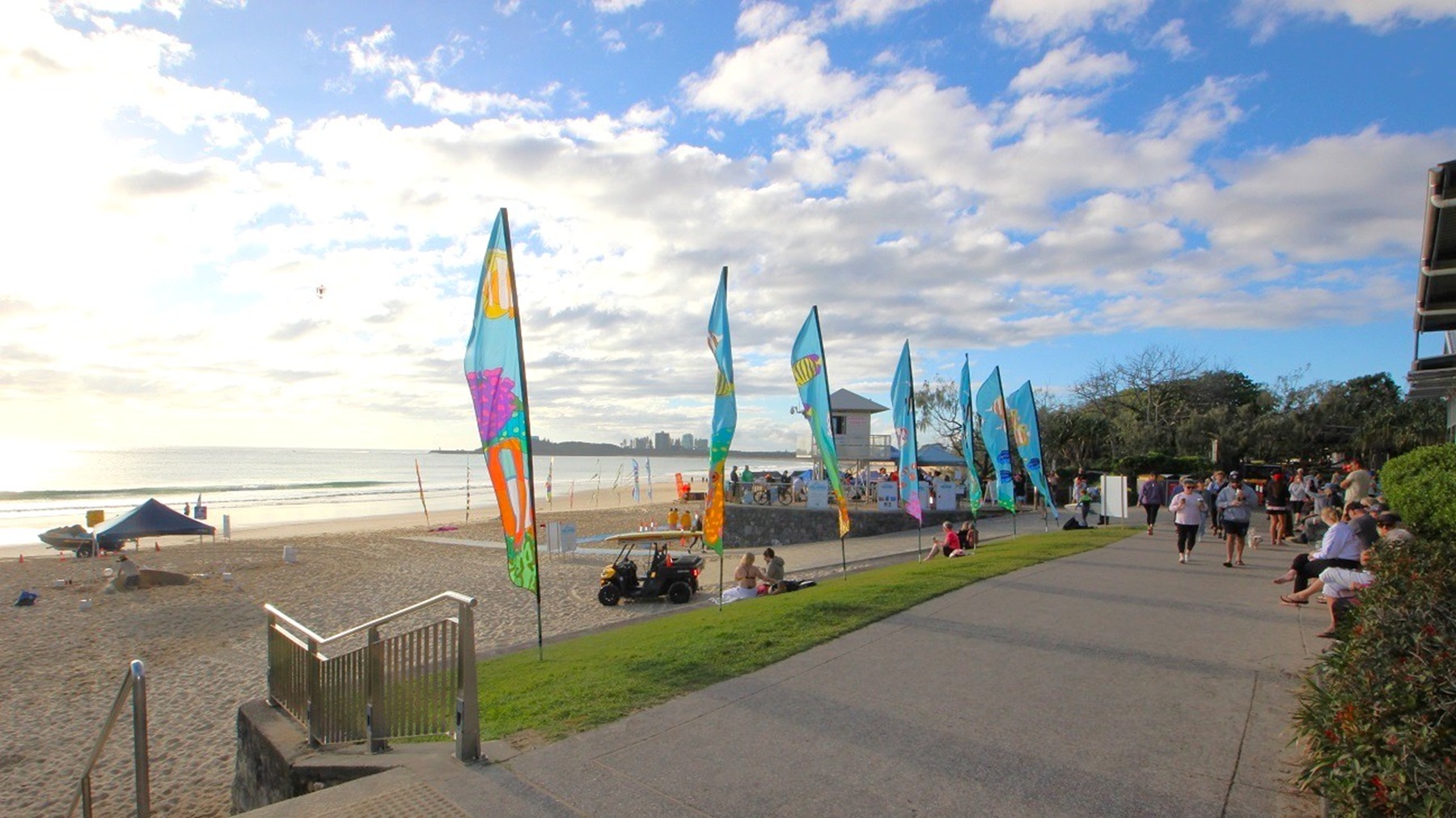 In the foreground, a paved pathway runs alongside the beach, with a strip of grass on the right. Colourful flags are planted along the pathway, creating a festive atmosphere. Just off the path, a small gazebo and several beach tents are visible, indicating an event setup. A few people are walking along the path, with others sitting on the grass. To the left, a set of steps leads down from the pathway to the sandy beach, which is dotted with beach equipment. In the foreground, a paved pathway runs alongside the beach, with a strip of grass on the right. Colourful flags are planted along the pathway, creating a festive atmosphere. Just off the path, a small gazebo and several beach tents are visible, indicating an event setup. A few people are walking along the path, with others sitting on the grass. To the left, a set of steps leads down from the pathway to the sandy beach, which is dotted with beach equipment.