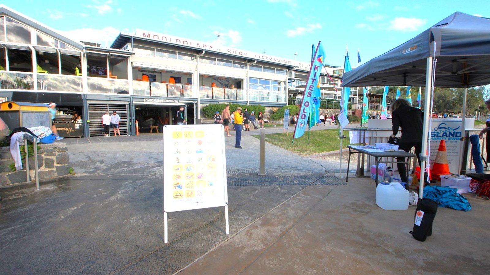 A pathway leads to a busy area, with a white sign displaying information prominently placed on the left. To the immediate right, a tent is set up with a table beneath it, where an individual is working, surrounded by various supplies. A gravel area is filled with people, with some seated at the adjacent Mooloolaba Surf Club, visible in the background. Flags promoting "Kites QLD" flutter alongside the path, while the upper level of the surf club features outdoor seating with bright orange chairs. A pathway leads to a busy area, with a white sign displaying information prominently placed on the left. To the immediate right, a tent is set up with a table beneath it, where an individual is working, surrounded by various supplies. A gravel area is filled with people, with some seated at the adjacent Mooloolaba Surf Club, visible in the background. Flags promoting "Kites QLD" flutter alongside the path, while the upper level of the surf club features outdoor seating with bright orange chairs.