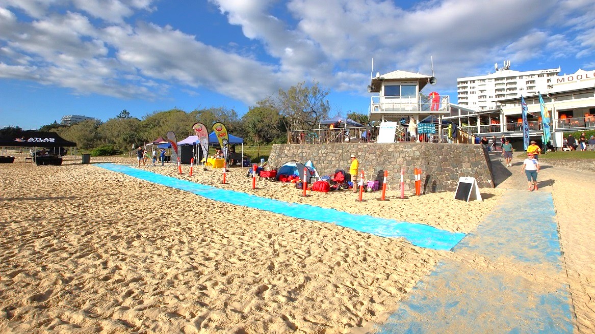 In the foreground, a sandy beach features a blue mat running towards the water, lined with orange cones. To the left, a black tent is set up, alongside several smaller tents, where event preparations are taking place. A short stone wall rises in the middle background, with a small lookout tower atop it, providing a vantage point for monitoring activities. The Mooloolaba Surf Club can be seen further back, with outdoor seating and people gathered on the upper level. In the foreground, a sandy beach features a blue mat running towards the water, lined with orange cones. To the left, a black tent is set up, alongside several smaller tents, where event preparations are taking place. A short stone wall rises in the middle background, with a small lookout tower atop it, providing a vantage point for monitoring activities. The Mooloolaba Surf Club can be seen further back, with outdoor seating and people gathered on the upper level.