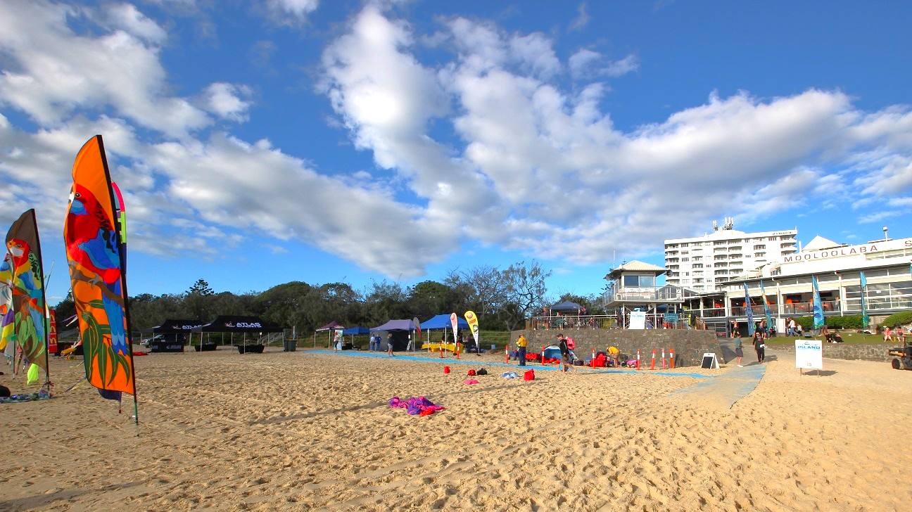 In the foreground, a sandy beach features colourful flags promoting various events, planted along the edge of a blue mat leading towards the water. To the left, a large black tent is set up, with a few people gathered nearby. Several smaller tents are located further along the beach area, where participants are preparing for activities. In the background, the multi-storey Mooloolaba Surf Club is clearly visible, with outdoor seating and a lookout tower. In the foreground, a sandy beach features colourful flags promoting various events, planted along the edge of a blue mat leading towards the water. To the left, a large black tent is set up, with a few people gathered nearby. Several smaller tents are located further along the beach area, where participants are preparing for activities. In the background, the multi-storey Mooloolaba Surf Club is clearly visible, with outdoor seating and a lookout tower.