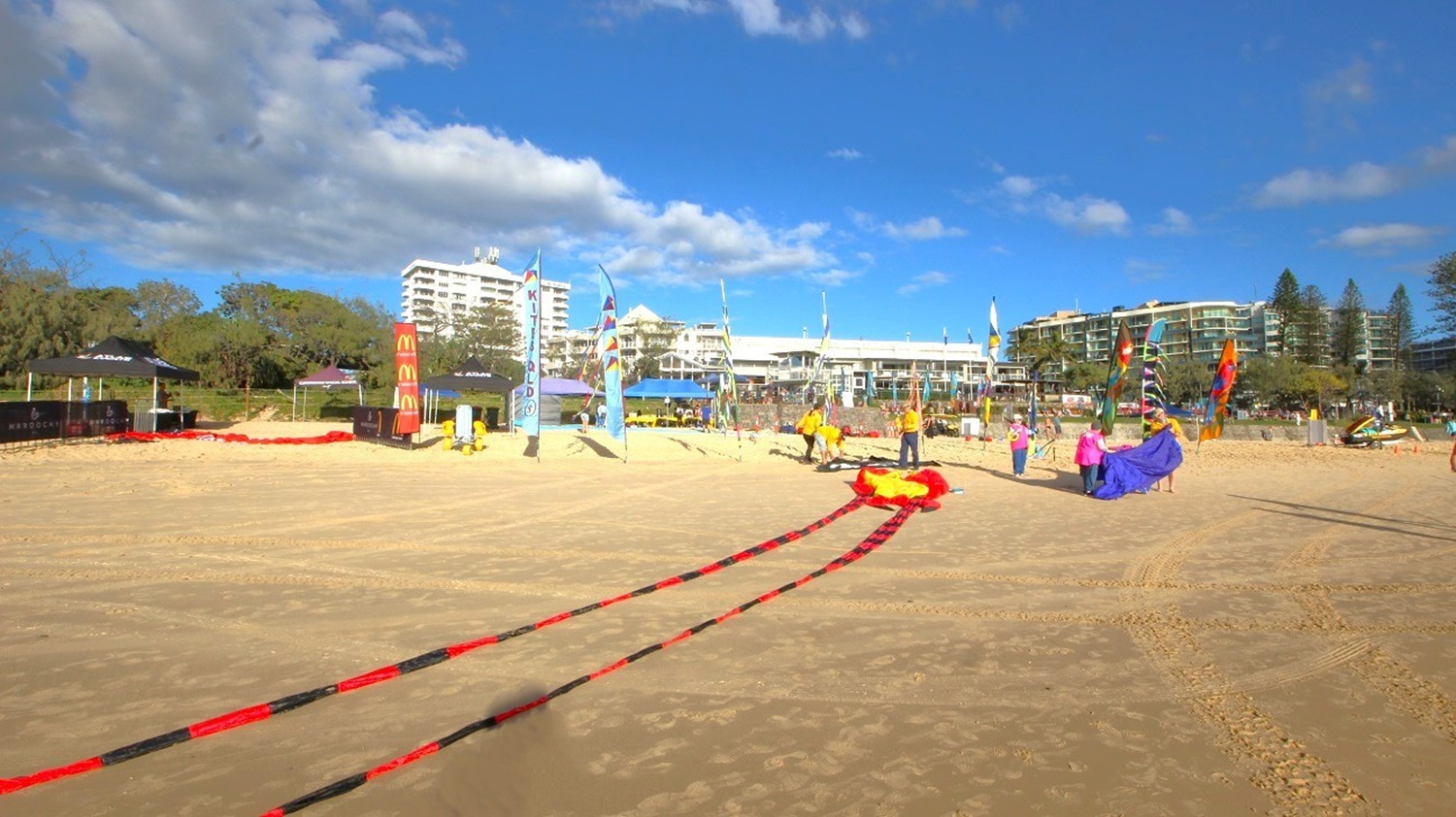 A sandy beach with a few people setting up colourful kites. In the background, there are several modern buildings and trees. Flags and banners are displayed near the beach, and a shadow of a person taking a photo is visible in the foreground. The sky is partly cloudy. A sandy beach with a few people setting up colourful kites. In the background, there are several modern buildings and trees. Flags and banners are displayed near the beach, and a shadow of a person taking a photo is visible in the foreground. The sky is partly cloudy.
