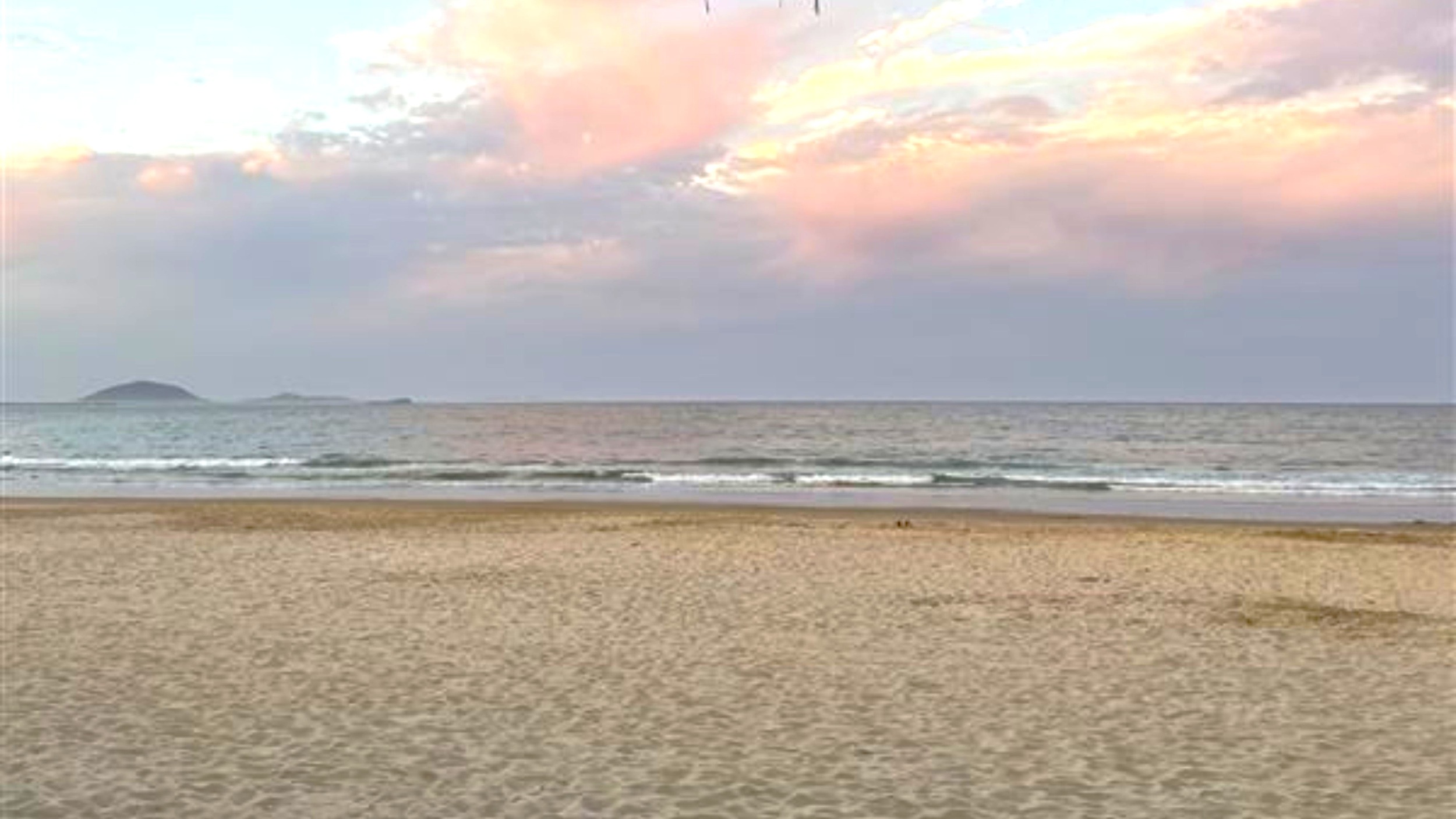 A sandy beach stretches across the foreground, leading to gentle waves lapping at the shore. In the background, the sea extends to the horizon, with small islands visible in the distance. The sky is filled with soft, pastel-coloured clouds, creating a serene atmosphere. A sandy beach stretches across the foreground, leading to gentle waves lapping at the shore. In the background, the sea extends to the horizon, with small islands visible in the distance. The sky is filled with soft, pastel-coloured clouds, creating a serene atmosphere.