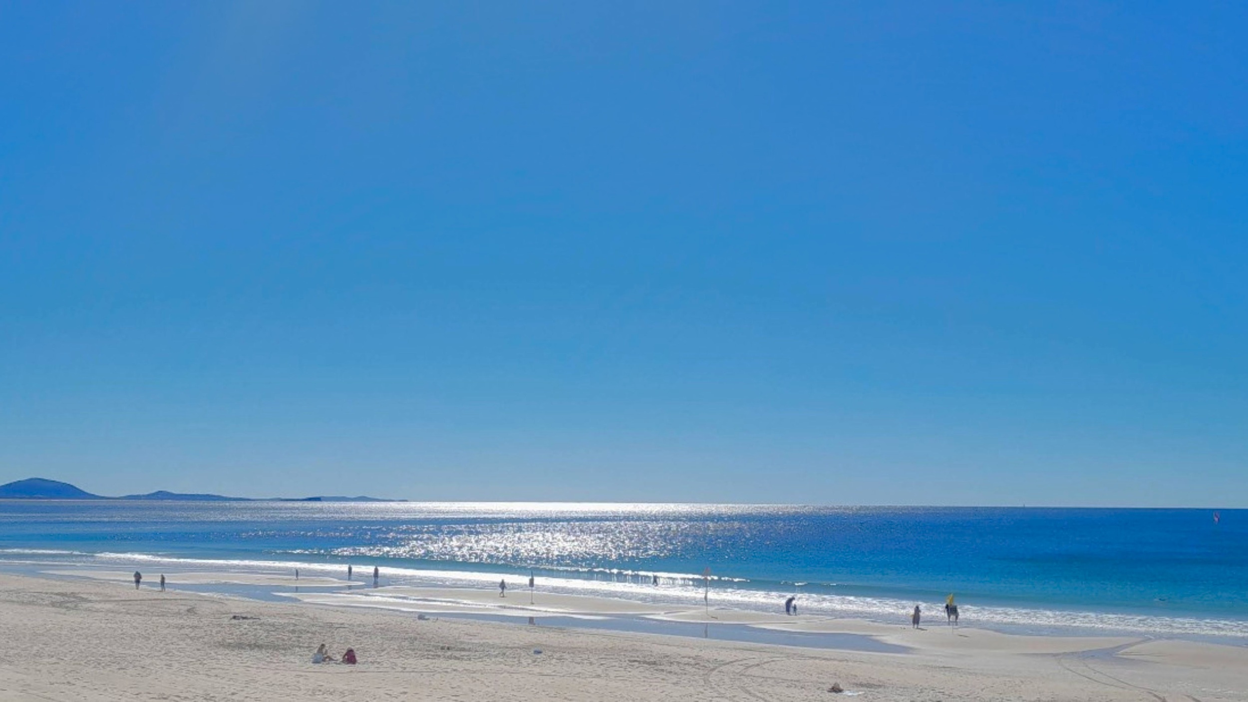 A sandy beach stretches across the foreground with a few people walking and sitting. The ocean is in the middle, with gentle waves and a shimmering surface reflecting the sunlight. In the background, low hills or islands are visible on the horizon. The sky is clear and blue. A sandy beach stretches across the foreground with a few people walking and sitting. The ocean is in the middle, with gentle waves and a shimmering surface reflecting the sunlight. In the background, low hills or islands are visible on the horizon. The sky is clear and blue.
