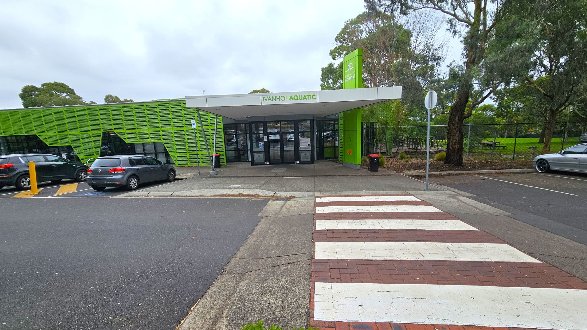 A building with a green facade and a sign reading "Ivanhoe Aquatic" above the entrance. The entrance has glass doors and a covered walkway. On the left, there are parked cars next to the building. On the right, there are trees and a grassy area with a picnic table. A zebra crossing leads to the entrance from the foreground, with a mix of asphalt and brick paving. A stop sign is visible near the trees. A building with a green facade and a sign reading "Ivanhoe Aquatic" above the entrance. The entrance has glass doors and a covered walkway. On the left, there are parked cars next to the building. On the right, there are trees and a grassy area with a picnic table. A zebra crossing leads to the entrance from the foreground, with a mix of asphalt and brick paving. A stop sign is visible near the trees.