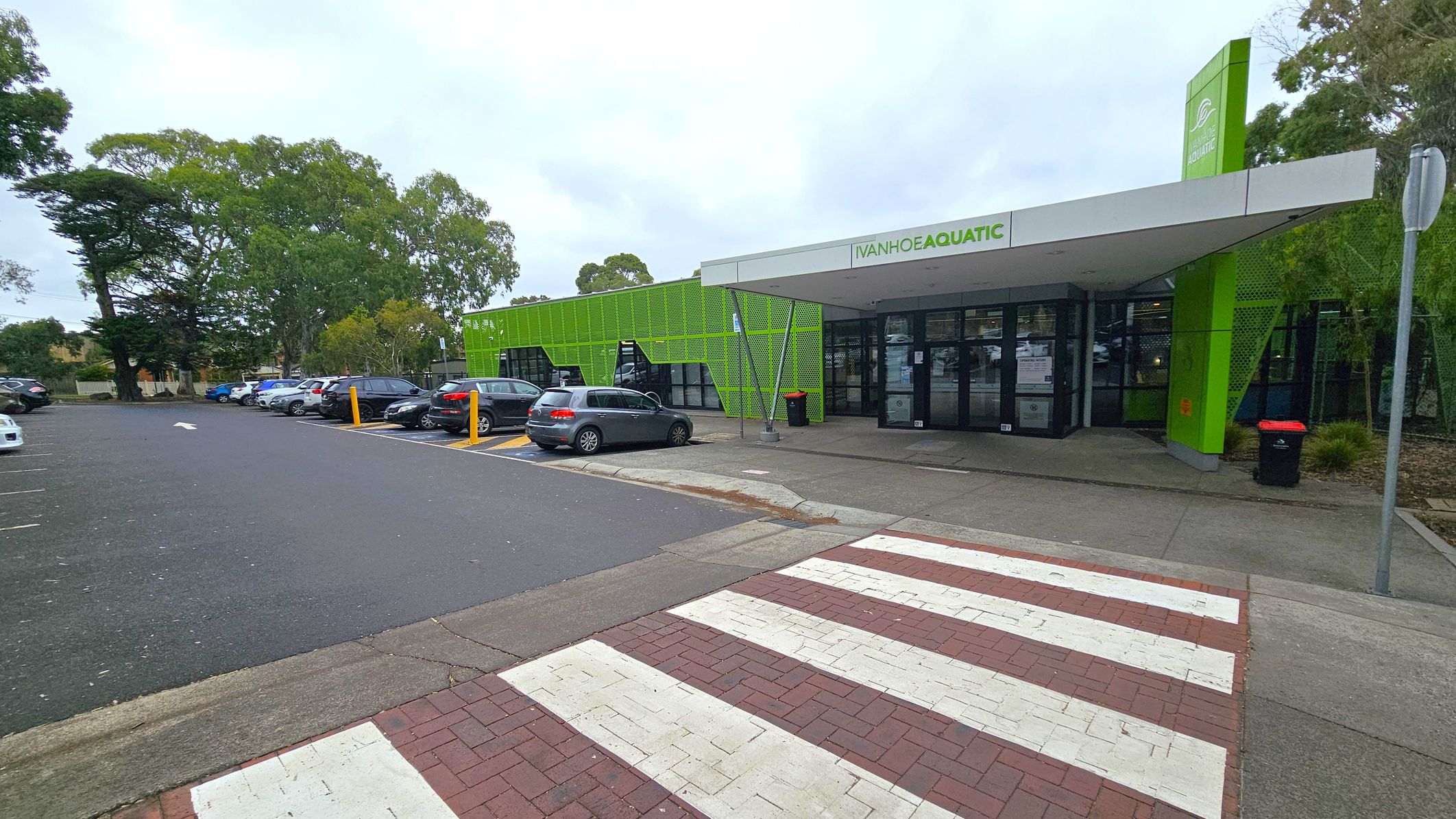 A modern building with a green facade and a sign reading "Ivanhoe Aquatic" above the entrance. The entrance has glass doors and a small awning. To the left, there is a car park with several parked cars and tall trees in the background. A zebra crossing leads to the entrance from the foreground, with a mix of red and white paving. A black bin is near the entrance, and a metal pole stands to the right. A modern building with a green facade and a sign reading "Ivanhoe Aquatic" above the entrance. The entrance has glass doors and a small awning. To the left, there is a car park with several parked cars and tall trees in the background. A zebra crossing leads to the entrance from the foreground, with a mix of red and white paving. A black bin is near the entrance, and a metal pole stands to the right.