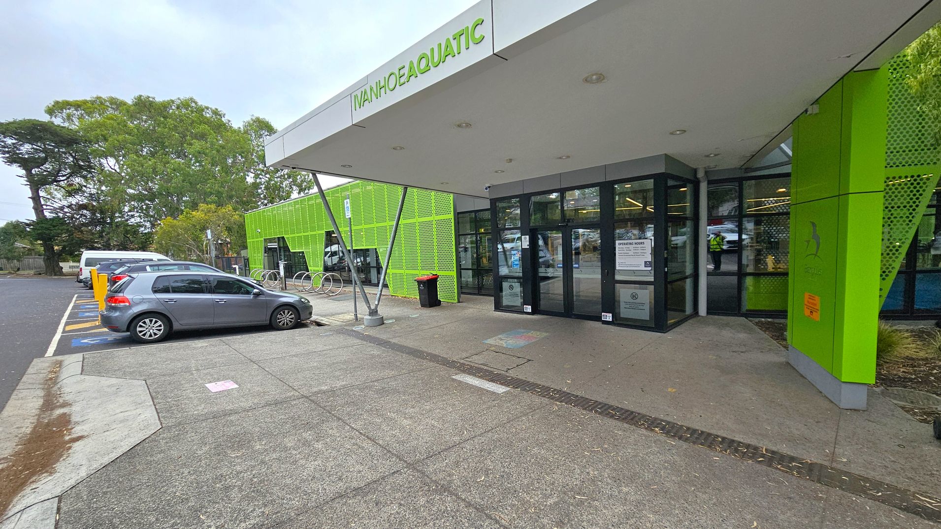 The image shows the entrance to a building with a sign reading "Ivanhoe Aquatic" above the door. The building has a modern design with bright green panels. On the left, there is a car park with a grey car and a white van parked in designated spaces. A row of bicycle racks is visible near the entrance. The ground is paved with concrete, and there are trees in the background. The entrance has glass doors with notices displayed on them. The image shows the entrance to a building with a sign reading "Ivanhoe Aquatic" above the door. The building has a modern design with bright green panels. On the left, there is a car park with a grey car and a white van parked in designated spaces. A row of bicycle racks is visible near the entrance. The ground is paved with concrete, and there are trees in the background. The entrance has glass doors with notices displayed on them.