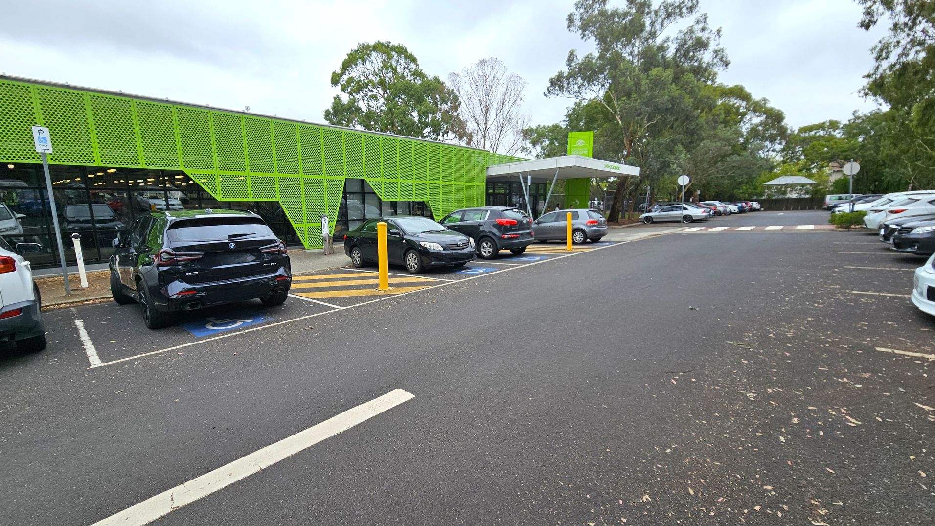 A car park with several vehicles is in front of a building with a distinctive green perforated facade on the left. The building has large glass windows beneath the green panels. The car park has designated spaces, including a disabled parking spot marked in blue. Yellow bollards are present near the entrance. Trees and foliage surround the area, with more parked cars visible in the background. The ground is asphalt, and there are some scattered leaves. A car park with several vehicles is in front of a building with a distinctive green perforated facade on the left. The building has large glass windows beneath the green panels. The car park has designated spaces, including a disabled parking spot marked in blue. Yellow bollards are present near the entrance. Trees and foliage surround the area, with more parked cars visible in the background. The ground is asphalt, and there are some scattered leaves.