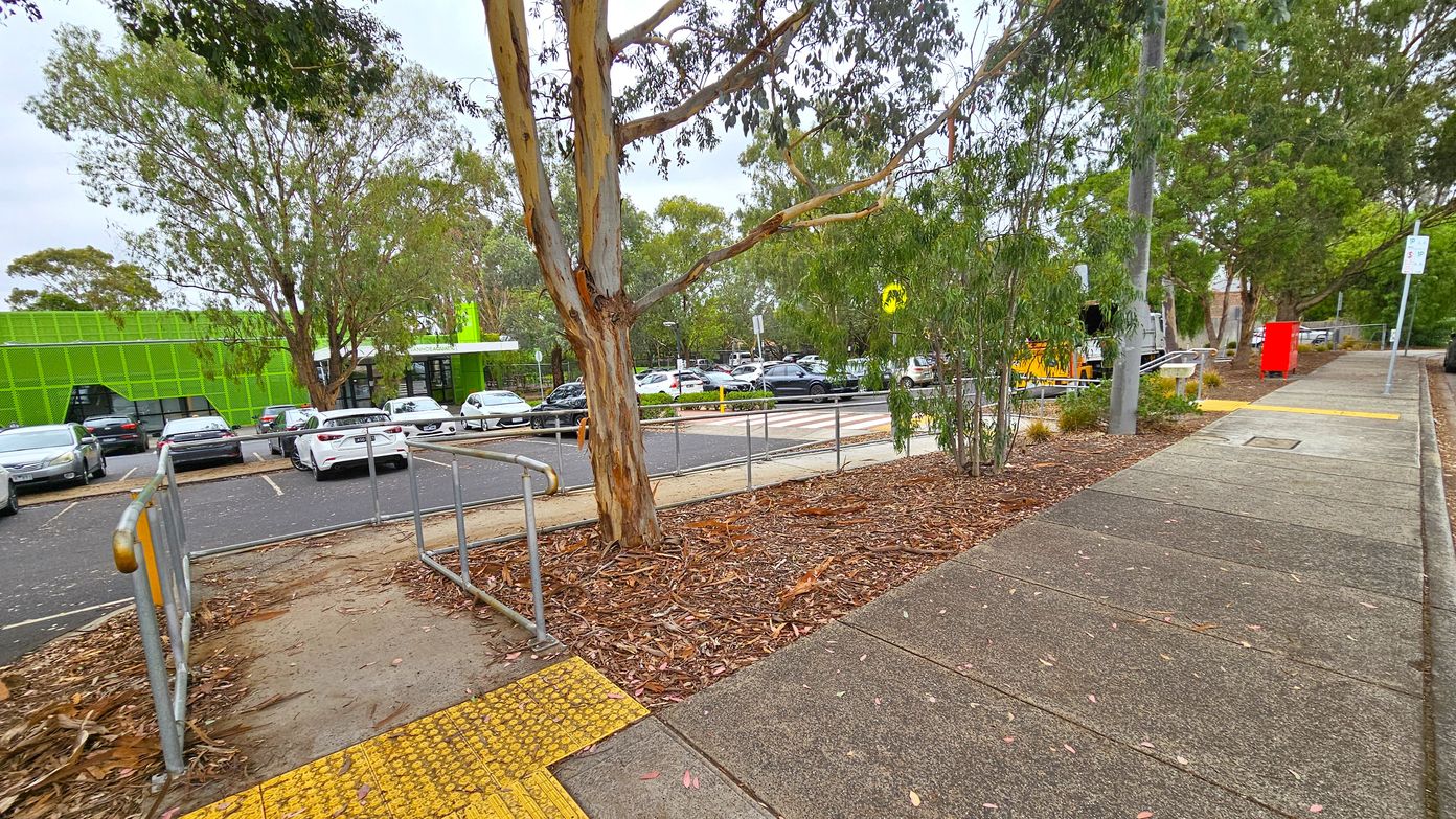 A footpath runs along the right side, bordered by a grassy area with trees. A bright green building is visible on the left, with a car park in front of it filled with vehicles. A large tree stands in the centre, surrounded by fallen leaves and bark. A metal railing lines a ramp leading from the footpath to the car park. A red post box is near the footpath, and a pedestrian crossing is visible in the background. A footpath runs along the right side, bordered by a grassy area with trees. A bright green building is visible on the left, with a car park in front of it filled with vehicles. A large tree stands in the centre, surrounded by fallen leaves and bark. A metal railing lines a ramp leading from the footpath to the car park. A red post box is near the footpath, and a pedestrian crossing is visible in the background.