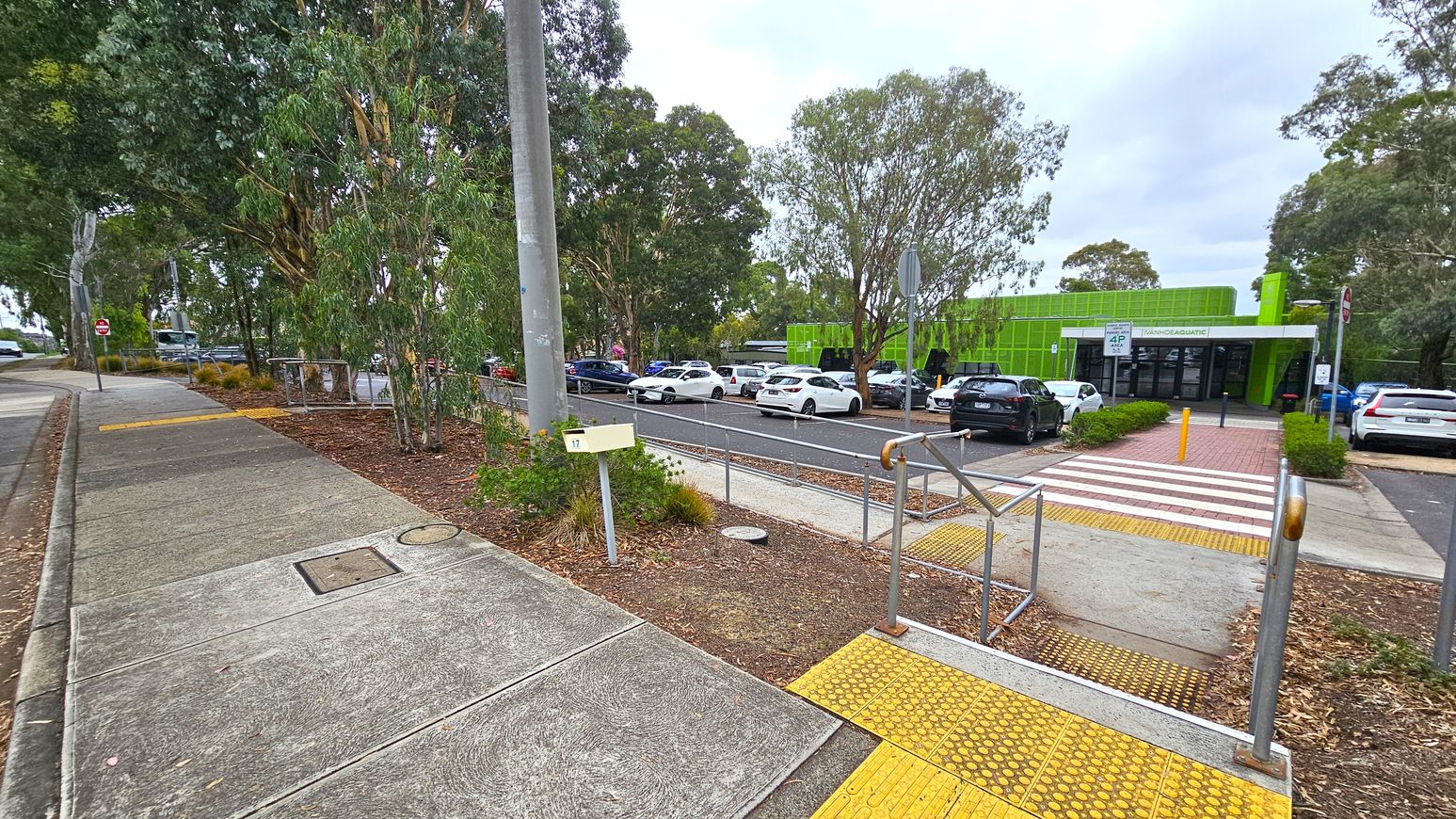 A footpath on the left with a concrete surface and a yellow tactile paving section leads to a road. On the right, a car park with several vehicles is visible, adjacent to a green building with a sign that reads "AQUATIC." Trees are scattered throughout the area, providing shade. A pedestrian crossing with red and white stripes connects the footpath to the building entrance. A metal railing and a utility pole are also present near the crossing. A footpath on the left with a concrete surface and a yellow tactile paving section leads to a road. On the right, a car park with several vehicles is visible, adjacent to a green building with a sign that reads "AQUATIC." Trees are scattered throughout the area, providing shade. A pedestrian crossing with red and white stripes connects the footpath to the building entrance. A metal railing and a utility pole are also present near the crossing.