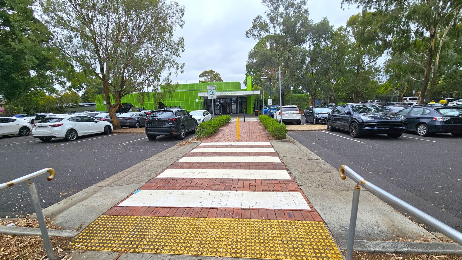 A pathway with a zebra crossing leads towards a building with a bright green facade in the centre. On either side of the path, there are parked cars, including white and dark-coloured vehicles. Trees with dense foliage are visible on both sides, providing shade. The ground is paved with bricks and asphalt, and there are metal railings on the left. The building has a sign above the entrance, and the area appears to be a car park. A pathway with a zebra crossing leads towards a building with a bright green facade in the centre. On either side of the path, there are parked cars, including white and dark-coloured vehicles. Trees with dense foliage are visible on both sides, providing shade. The ground is paved with bricks and asphalt, and there are metal railings on the left. The building has a sign above the entrance, and the area appears to be a car park.