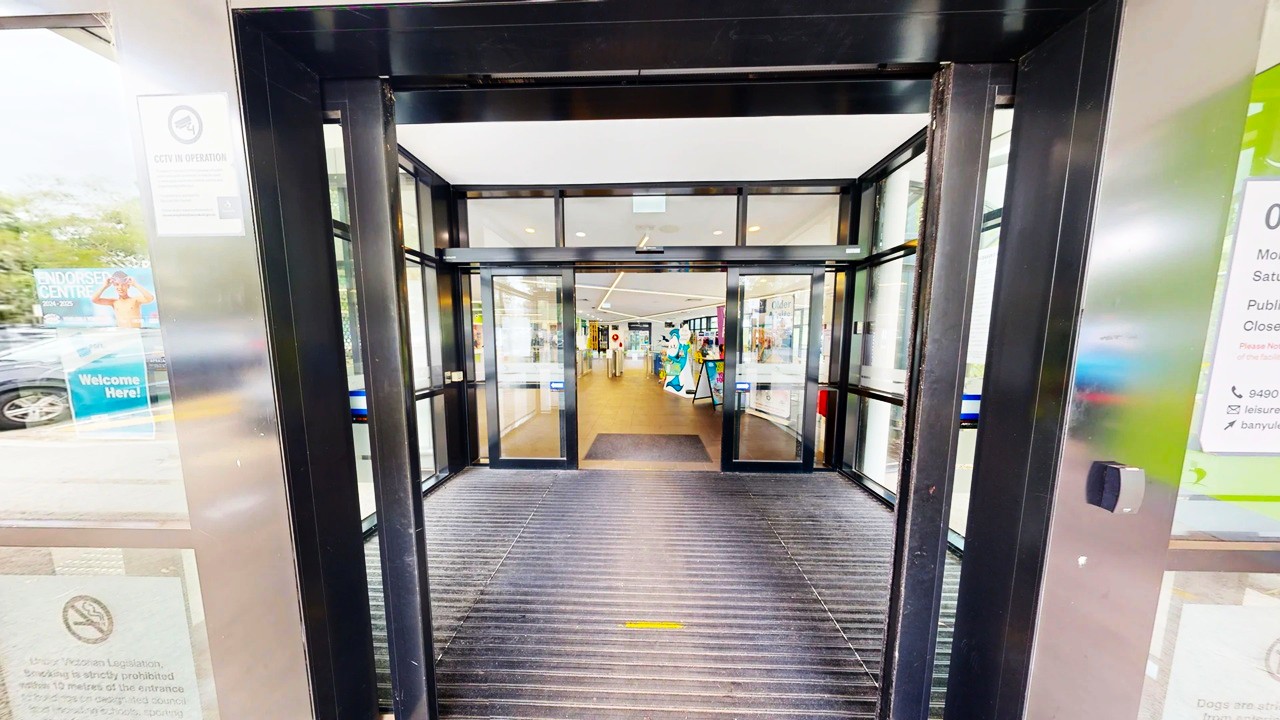 Automatic sliding glass doors lead into a building with a bright interior. The entrance has a black frame and a ribbed mat on the floor. Inside, the floor is wooden, and there are displays and signs visible in the distance. On the left, there are posters and signs on the glass, while the right side also features signage. The ceiling inside is white with recessed lighting. Automatic sliding glass doors lead into a building with a bright interior. The entrance has a black frame and a ribbed mat on the floor. Inside, the floor is wooden, and there are displays and signs visible in the distance. On the left, there are posters and signs on the glass, while the right side also features signage. The ceiling inside is white with recessed lighting.