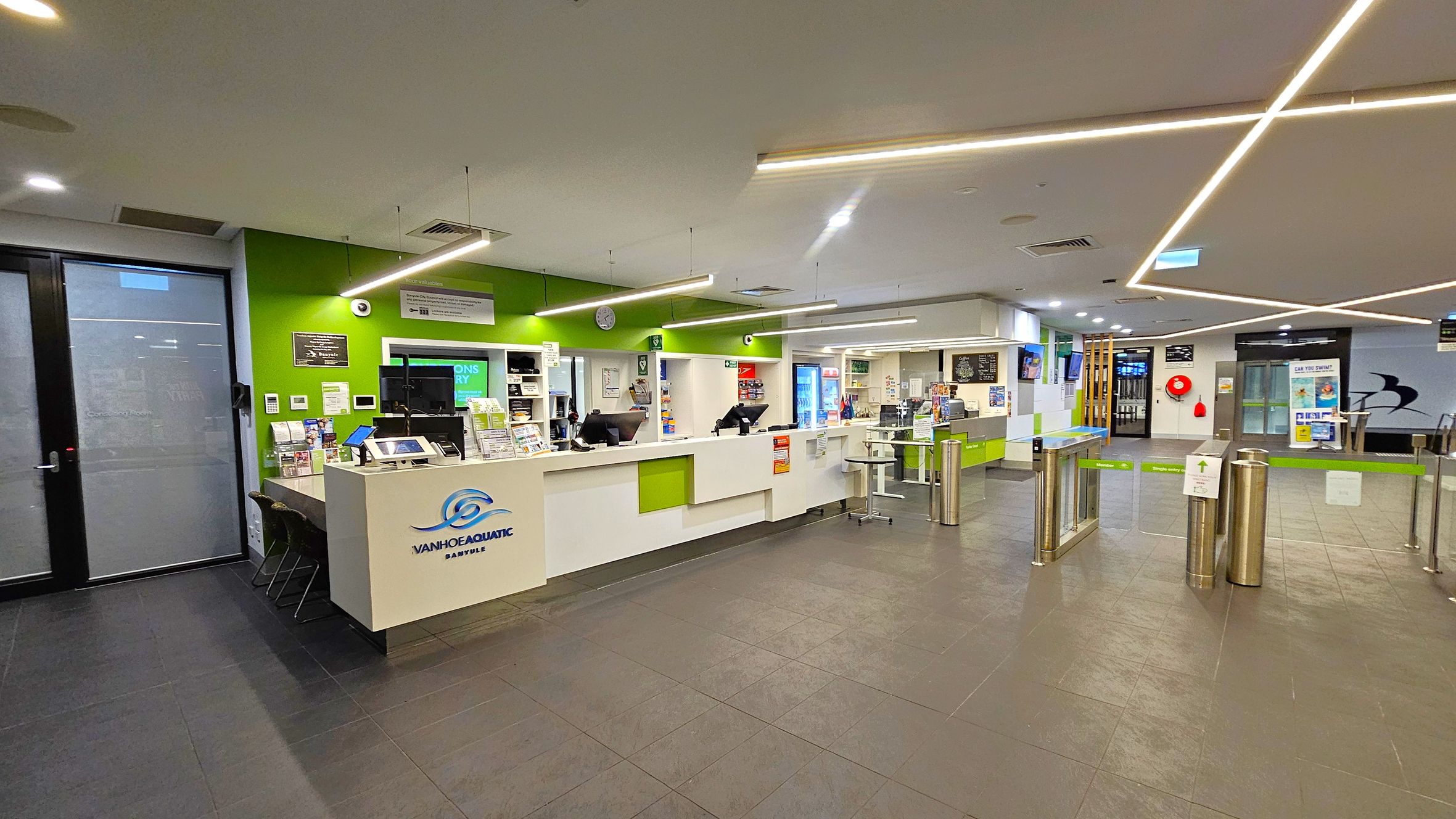 A modern reception area with a green and white colour scheme. On the left, there's a counter with brochures and a computer, and a door with frosted glass. In the centre, the reception desk displays the "Ivanhoe Aquatic" logo, with computers and informational materials. On the right, there are turnstiles leading to another area. The ceiling features linear lights, and the floor is tiled in dark grey. Various signs and posters are visible throughout the space. A modern reception area with a green and white colour scheme. On the left, there's a counter with brochures and a computer, and a door with frosted glass. In the centre, the reception desk displays the "Ivanhoe Aquatic" logo, with computers and informational materials. On the right, there are turnstiles leading to another area. The ceiling features linear lights, and the floor is tiled in dark grey. Various signs and posters are visible throughout the space.