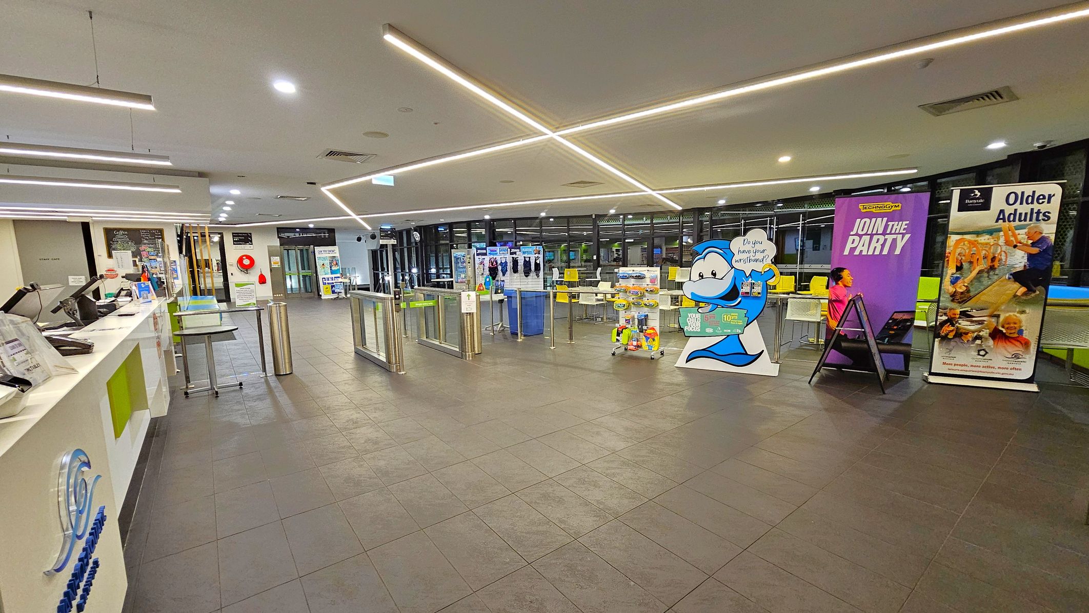 A spacious indoor area with a tiled floor and a ceiling featuring linear lights. On the left, there's a reception counter with computers and informational materials. In the centre, there are turnstiles leading to a seating area with tables and chairs. On the right, several promotional banners are displayed, including one with a cartoon character and others advertising activities for adults. The background has large windows and additional seating. A spacious indoor area with a tiled floor and a ceiling featuring linear lights. On the left, there's a reception counter with computers and informational materials. In the centre, there are turnstiles leading to a seating area with tables and chairs. On the right, several promotional banners are displayed, including one with a cartoon character and others advertising activities for adults. The background has large windows and additional seating.