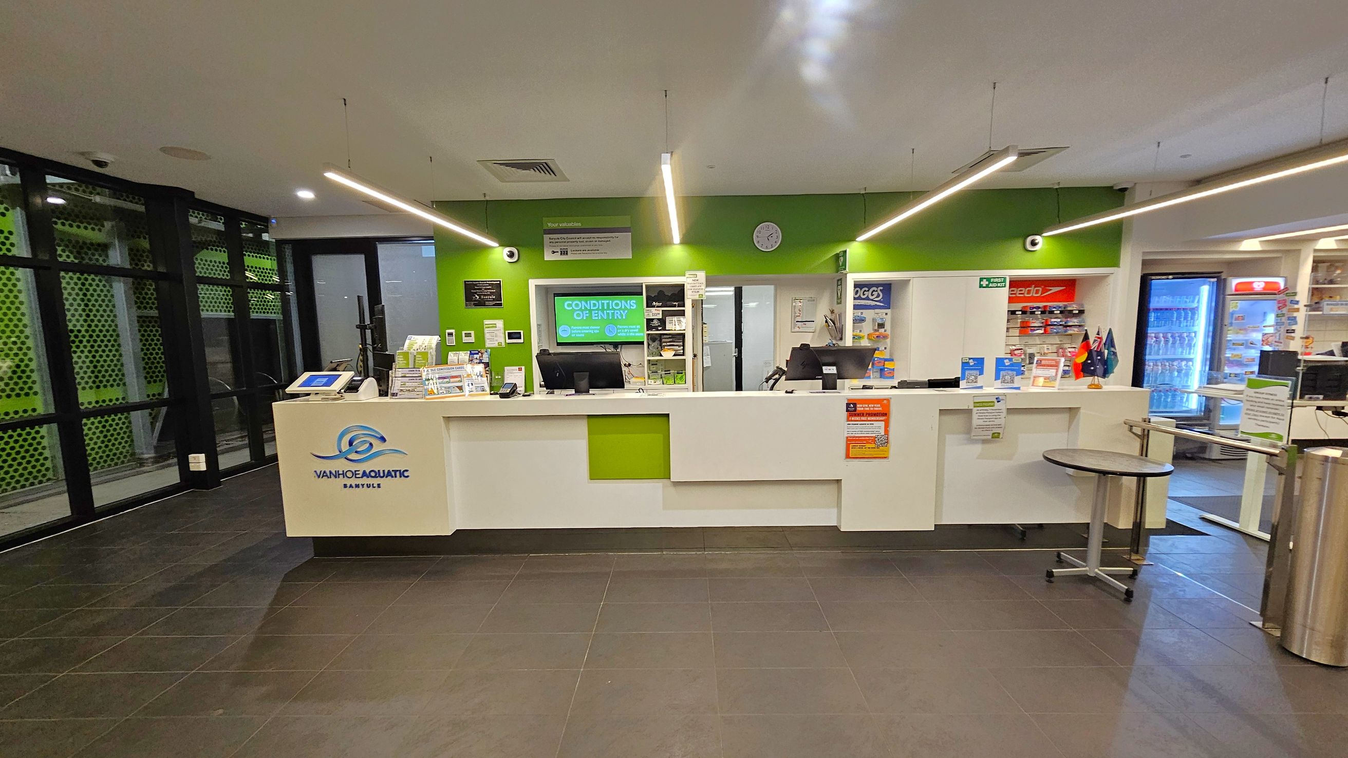 A reception area with a white counter featuring a green accent and a logo. Behind the counter, a green wall displays a clock and various signs. To the left, there are large windows with a perforated green pattern. On the right, shelves with products and a refrigerator with drinks are visible. The floor is tiled in dark grey, and the ceiling has linear lights. A small round table is in front of the counter. A reception area with a white counter featuring a green accent and a logo. Behind the counter, a green wall displays a clock and various signs. To the left, there are large windows with a perforated green pattern. On the right, shelves with products and a refrigerator with drinks are visible. The floor is tiled in dark grey, and the ceiling has linear lights. A small round table is in front of the counter.