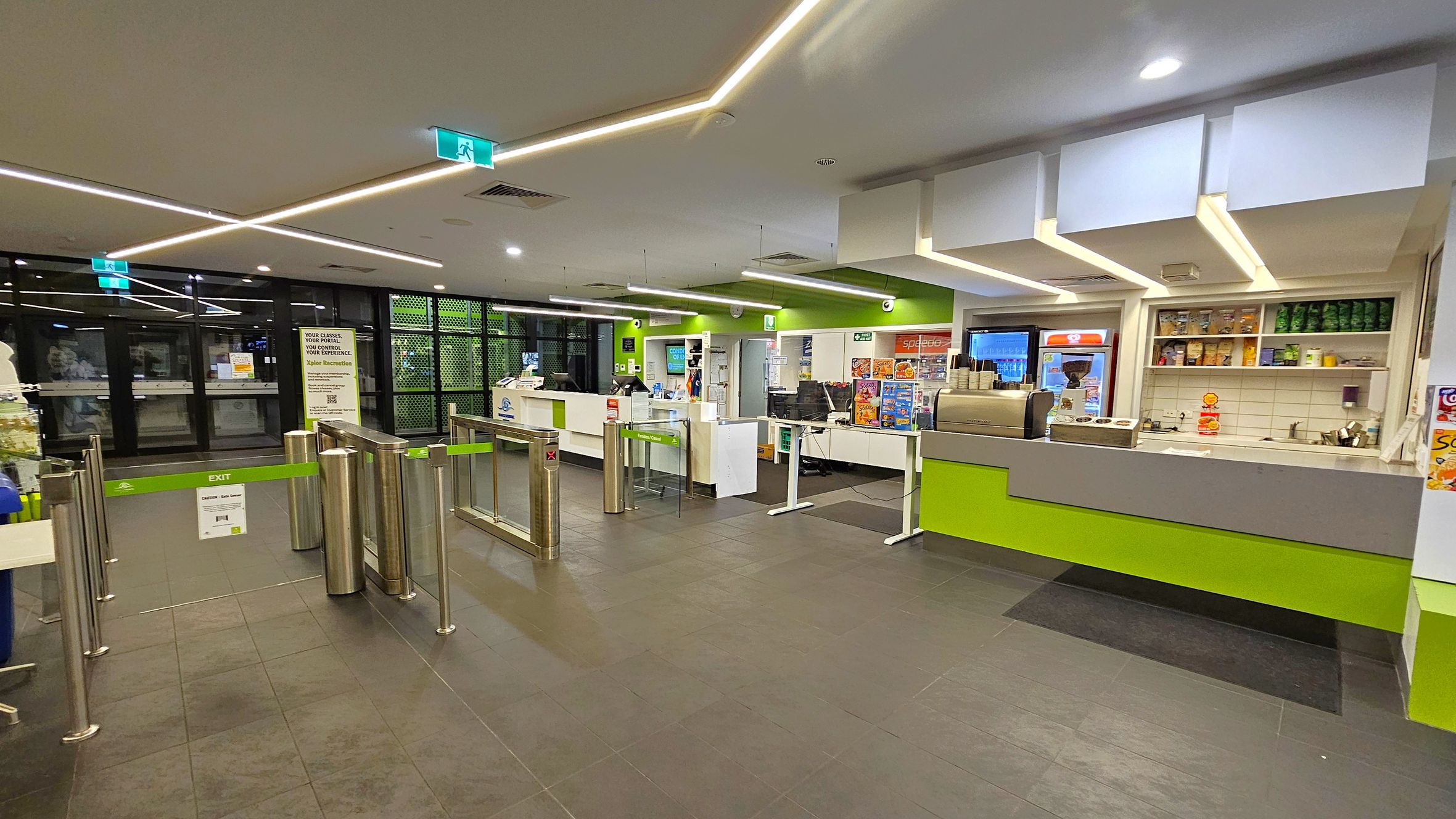 A modern indoor space with a tiled floor and a ceiling featuring linear lights. On the left, there are glass doors with a green exit sign above and a turnstile gate with a green exit barrier. In the centre, a counter with various items and a sign. On the right, a service counter with a coffee machine, shelves stocked with snacks, and a menu board. The walls are painted green and white, and the area is well-lit. A modern indoor space with a tiled floor and a ceiling featuring linear lights. On the left, there are glass doors with a green exit sign above and a turnstile gate with a green exit barrier. In the centre, a counter with various items and a sign. On the right, a service counter with a coffee machine, shelves stocked with snacks, and a menu board. The walls are painted green and white, and the area is well-lit.