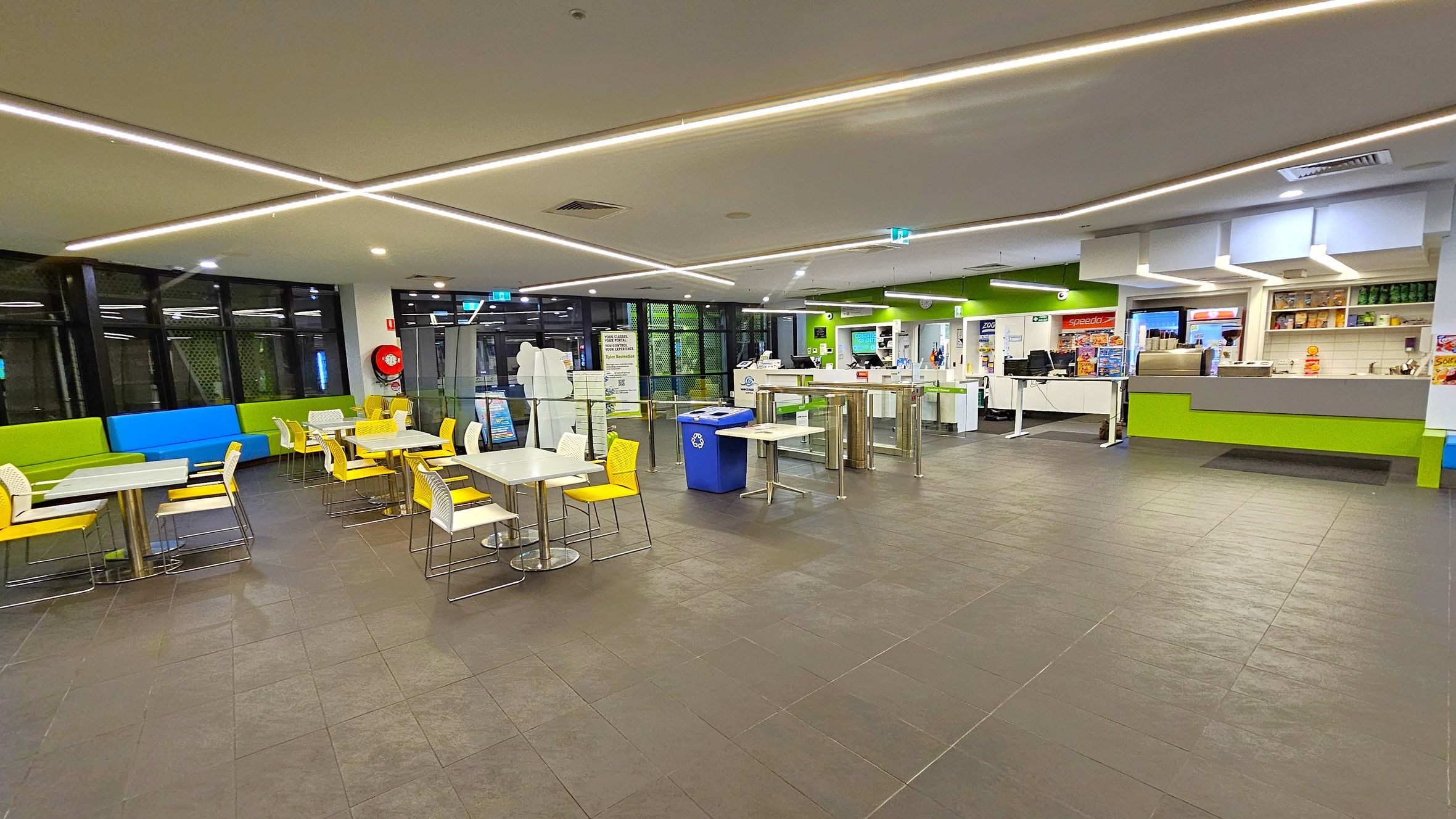 A spacious indoor cafeteria with a tiled floor and a ceiling featuring linear lights. On the left, there are tables with white tops and yellow chairs, alongside green and blue cushioned bench seating. In the centre, a blue recycling bin is visible. On the right, a counter area with green accents displays various items, with shelves holding products above. The background includes large windows and a fire extinguisher mounted on the wall. A spacious indoor cafeteria with a tiled floor and a ceiling featuring linear lights. On the left, there are tables with white tops and yellow chairs, alongside green and blue cushioned bench seating. In the centre, a blue recycling bin is visible. On the right, a counter area with green accents displays various items, with shelves holding products above. The background includes large windows and a fire extinguisher mounted on the wall.