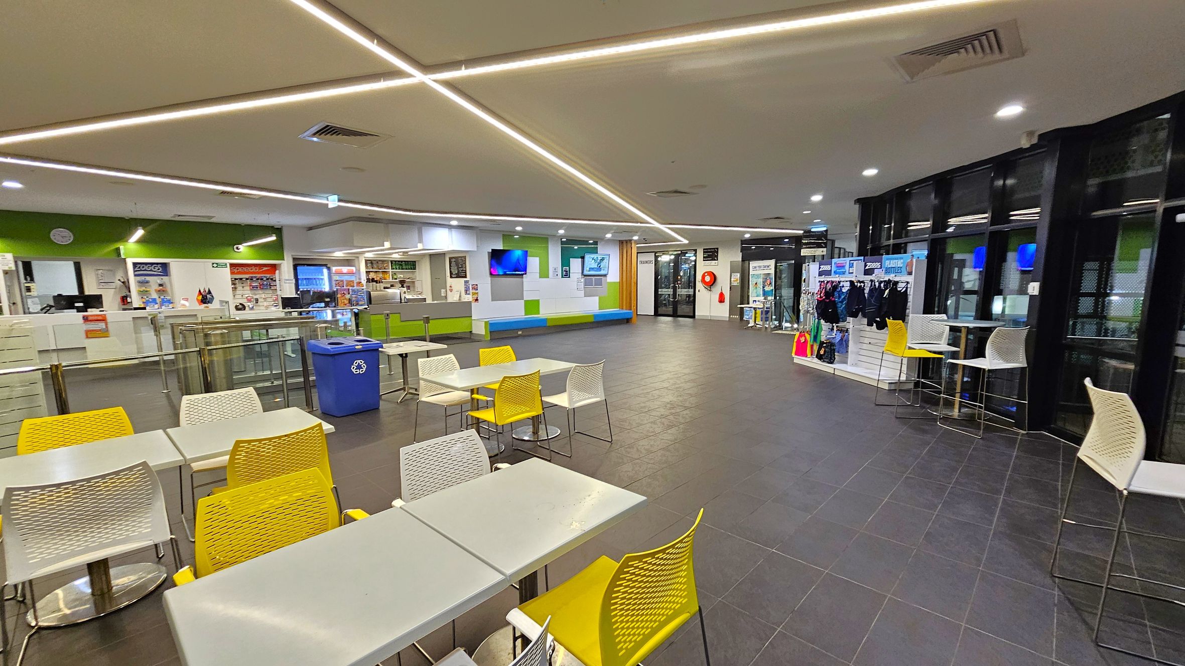 A spacious indoor area with a tiled floor and a ceiling featuring linear lights. On the left, there are tables with yellow and white chairs, and a blue recycling bin. The background has a counter with a green accent wall, a clock, and a display of products. In the centre, a wall-mounted TV and a seating area with green and blue cushions are visible. On the right, there is a display of swimwear and accessories, with high tables and chairs near large windows. A spacious indoor area with a tiled floor and a ceiling featuring linear lights. On the left, there are tables with yellow and white chairs, and a blue recycling bin. The background has a counter with a green accent wall, a clock, and a display of products. In the centre, a wall-mounted TV and a seating area with green and blue cushions are visible. On the right, there is a display of swimwear and accessories, with high tables and chairs near large windows.