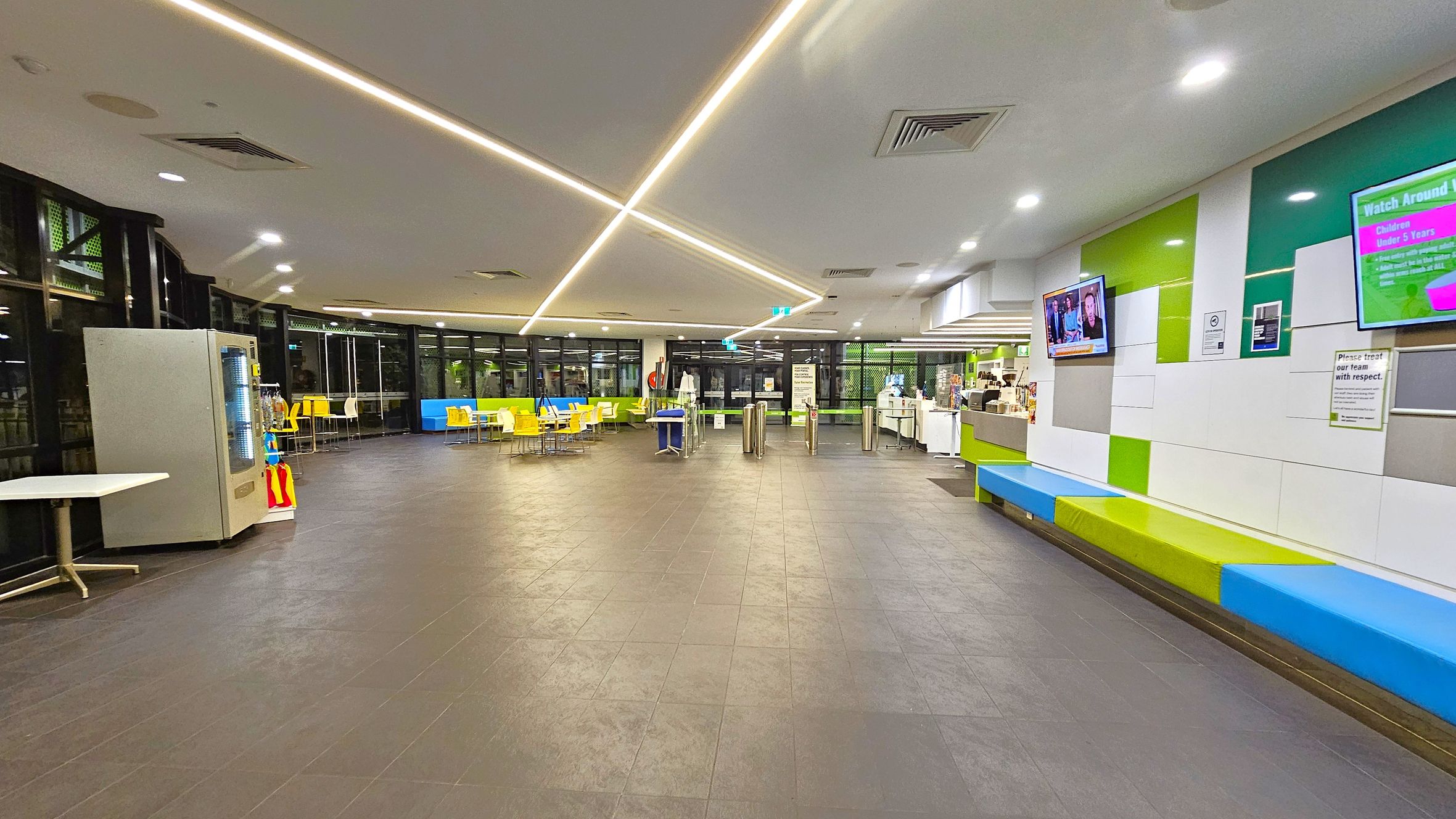 A spacious indoor area with a dark tiled floor and a ceiling featuring recessed lighting and linear light strips. On the left, there's a vending machine and a small table. In the centre, yellow chairs and tables are arranged near large windows. On the right, a wall with colourful panels has two mounted screens and a long bench with blue and green cushions. A counter with various items is visible further back on the right. A spacious indoor area with a dark tiled floor and a ceiling featuring recessed lighting and linear light strips. On the left, there's a vending machine and a small table. In the centre, yellow chairs and tables are arranged near large windows. On the right, a wall with colourful panels has two mounted screens and a long bench with blue and green cushions. A counter with various items is visible further back on the right.