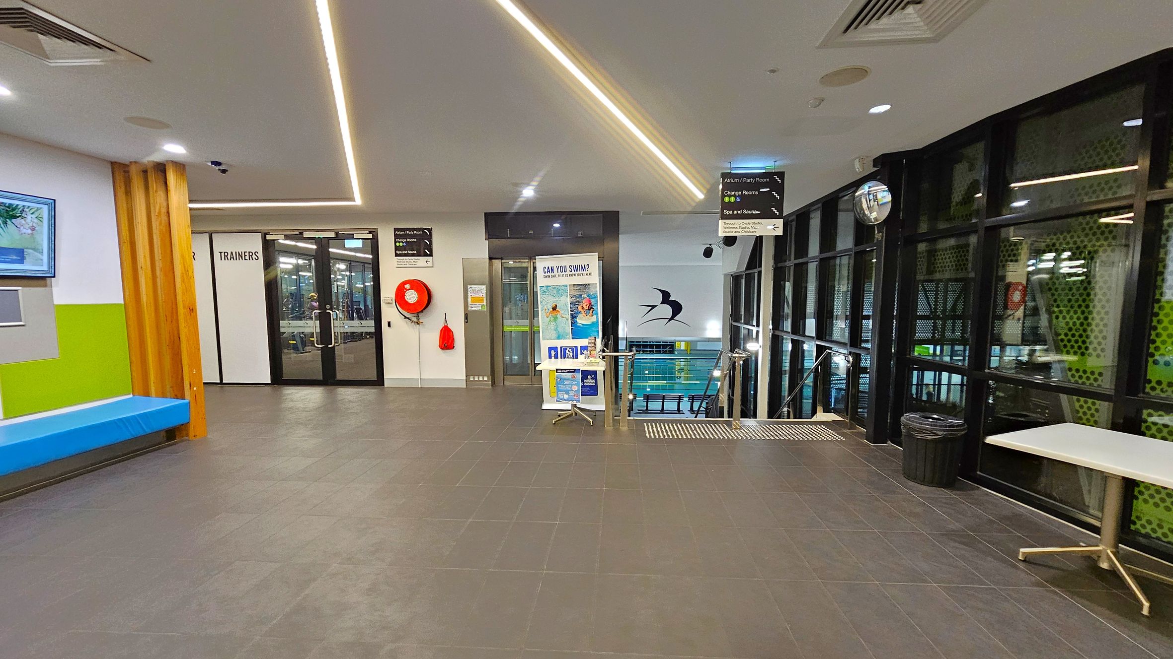 A modern indoor facility with a grey tiled floor and a white ceiling featuring recessed lighting. On the left, there's a blue bench against a wall with green and white sections, and a digital screen above. In the centre, glass doors labelled "TRAINERS" lead to another room. A red fire hose reel is mounted on the wall. To the right, large windows overlook a pool area, with a table and a bin nearby. A sign above directs to various amenities, and a small table with posters stands near the pool entrance. A modern indoor facility with a grey tiled floor and a white ceiling featuring recessed lighting. On the left, there's a blue bench against a wall with green and white sections, and a digital screen above. In the centre, glass doors labelled "TRAINERS" lead to another room. A red fire hose reel is mounted on the wall. To the right, large windows overlook a pool area, with a table and a bin nearby. A sign above directs to various amenities, and a small table with posters stands near the pool entrance.