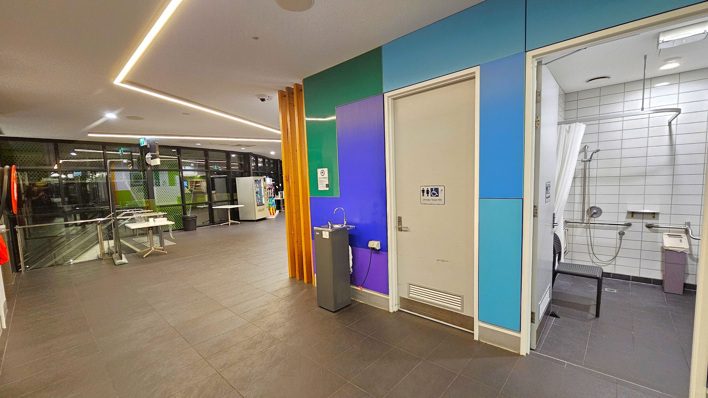 A modern indoor space with a tiled floor and a ceiling featuring recessed lighting. On the right, there is an accessible bathroom with white tiled walls, a shower, and a chair. The door is surrounded by panels in shades of blue and green. A drinking fountain is positioned next to the door. On the left, large windows with a view of the outside are visible, along with a vending machine and tables. A life preserver is mounted on the wall near the windows. A modern indoor space with a tiled floor and a ceiling featuring recessed lighting. On the right, there is an accessible bathroom with white tiled walls, a shower, and a chair. The door is surrounded by panels in shades of blue and green. A drinking fountain is positioned next to the door. On the left, large windows with a view of the outside are visible, along with a vending machine and tables. A life preserver is mounted on the wall near the windows.