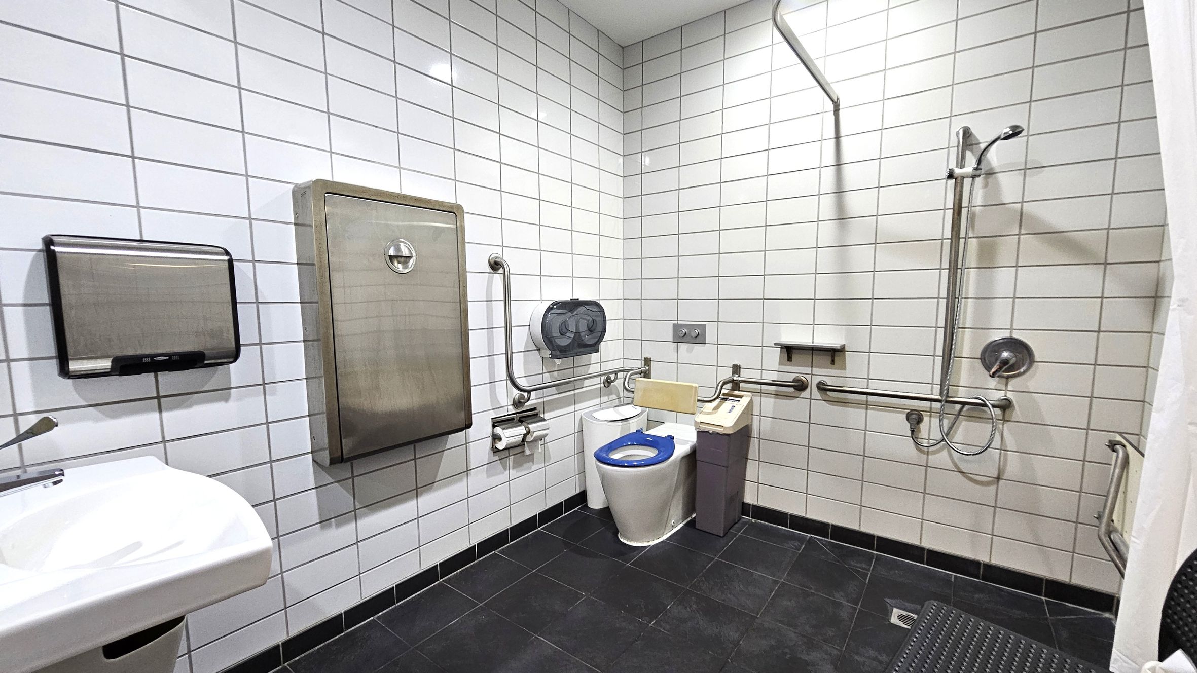 A bathroom with white tiled walls and a dark tiled floor. On the left, there's a white sink with a soap dispenser and a paper towel holder above it. In the centre, a toilet with a blue seat is equipped with support rails and a sanitary bin nearby. On the right, a shower area features a handheld showerhead and additional support rails. A large stainless steel panel is mounted on the wall above the toilet. The ceiling is plain and white. A bathroom with white tiled walls and a dark tiled floor. On the left, there's a white sink with a soap dispenser and a paper towel holder above it. In the centre, a toilet with a blue seat is equipped with support rails and a sanitary bin nearby. On the right, a shower area features a handheld showerhead and additional support rails. A large stainless steel panel is mounted on the wall above the toilet. The ceiling is plain and white.