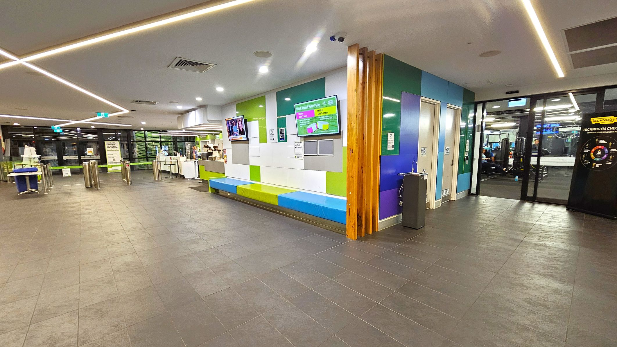 A modern interior with a tiled floor and a ceiling featuring recessed lighting. On the right, there is a colourful wall with a bench and digital screens displaying information. A water fountain is nearby. On the left, there are turnstiles and a reception area with a counter. Glass doors lead to a gym area with exercise equipment visible. The space is well-lit and has a contemporary design. A modern interior with a tiled floor and a ceiling featuring recessed lighting. On the right, there is a colourful wall with a bench and digital screens displaying information. A water fountain is nearby. On the left, there are turnstiles and a reception area with a counter. Glass doors lead to a gym area with exercise equipment visible. The space is well-lit and has a contemporary design.