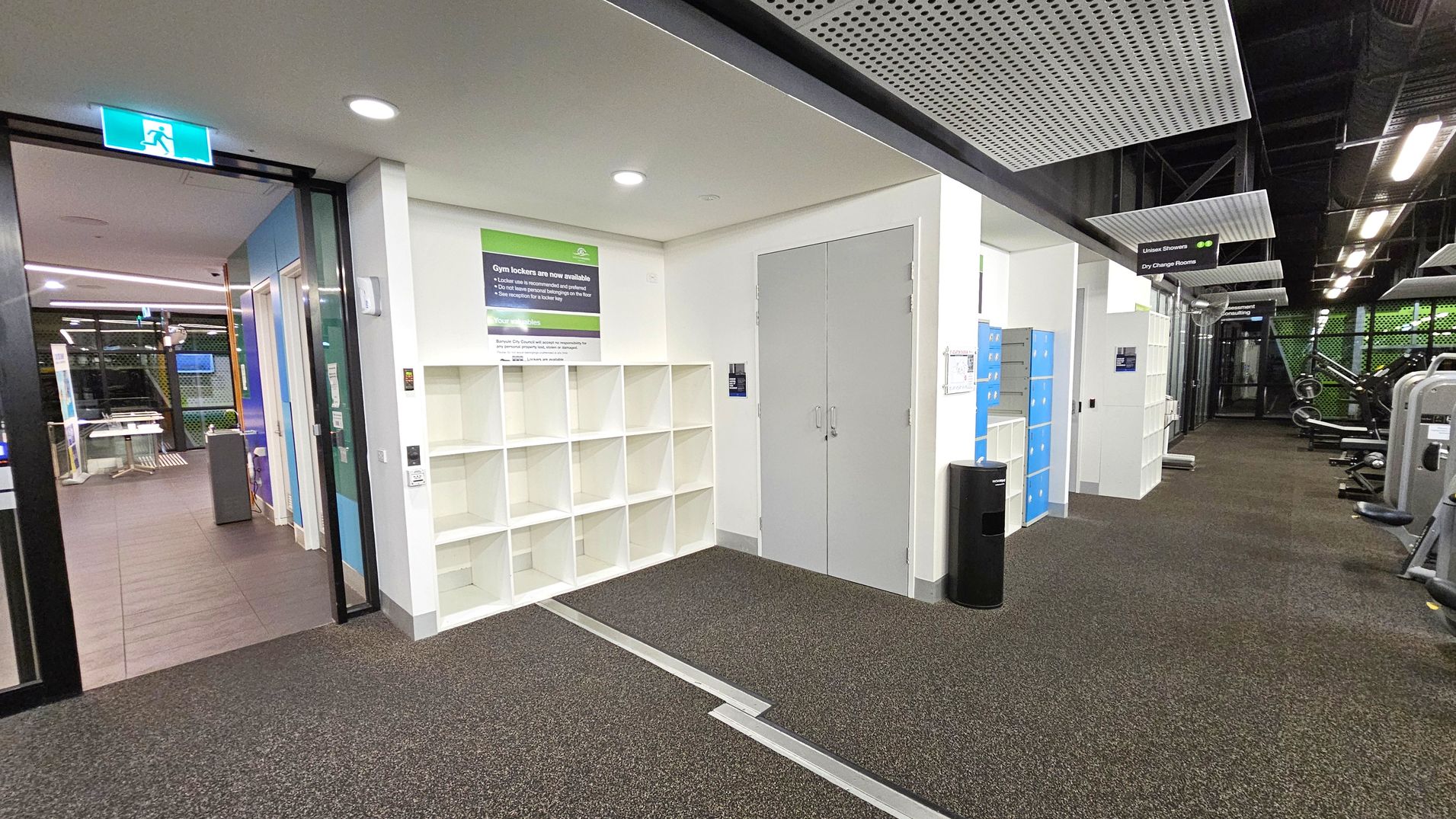 On the left, there's a glass door with an illuminated exit sign above it, leading to a room with tables and chairs. In the centre, white cubby shelves are mounted on the wall beneath a sign about gym lockers. To the right, blue lockers and gym equipment are visible. The floor is carpeted in a speckled pattern, and the ceiling features perforated panels with exposed ductwork. On the left, there's a glass door with an illuminated exit sign above it, leading to a room with tables and chairs. In the centre, white cubby shelves are mounted on the wall beneath a sign about gym lockers. To the right, blue lockers and gym equipment are visible. The floor is carpeted in a speckled pattern, and the ceiling features perforated panels with exposed ductwork.