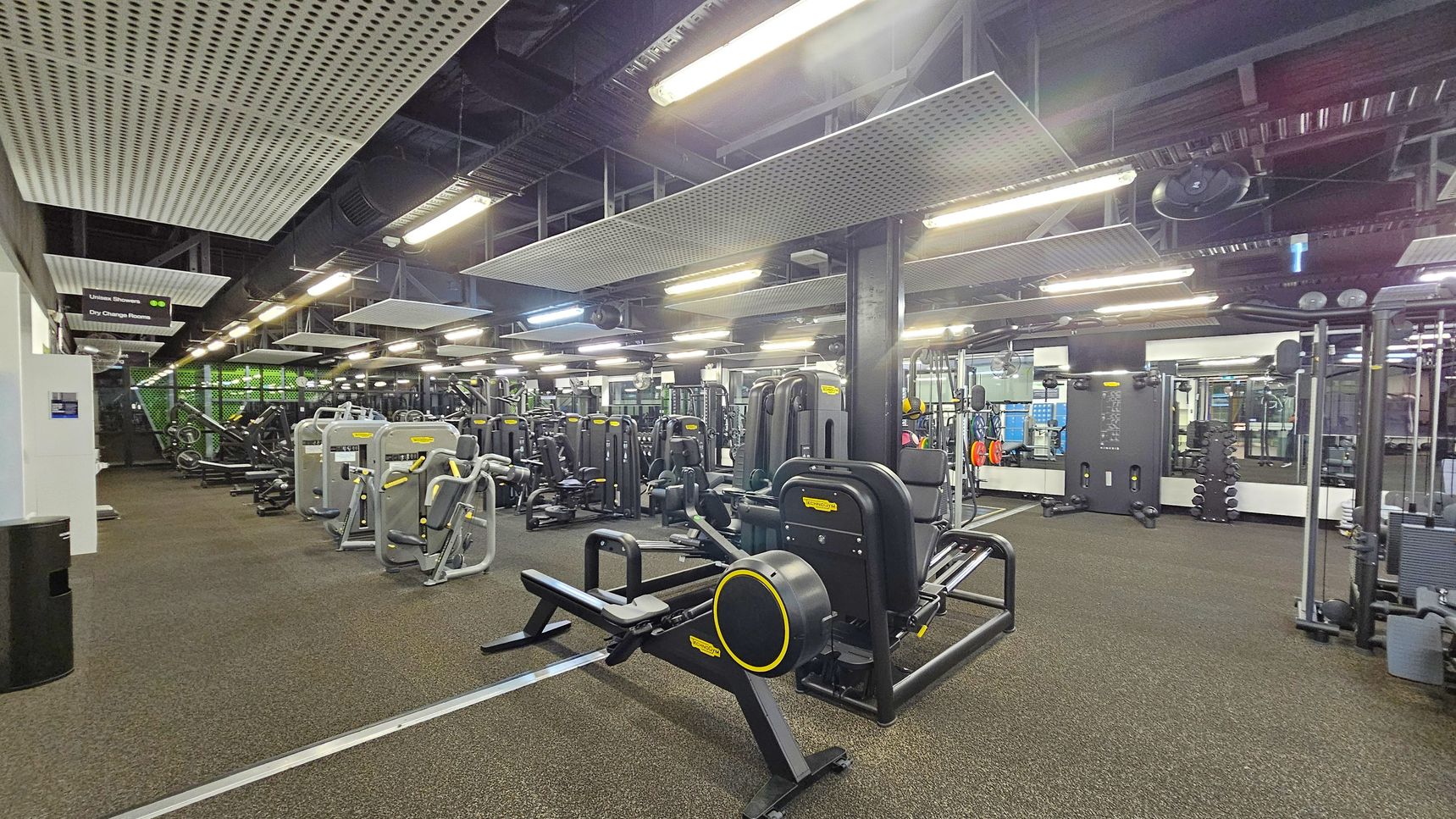 A spacious gym interior with various exercise machines arranged in rows. On the left, there are signs for union showers and dry change rooms. The floor is covered with a speckled rubber mat, and the ceiling features large, rectangular acoustic panels and fluorescent lights. In the centre, there are multiple strength training machines, and on the right, a wall with mirrors and additional equipment, including weights and resistance machines. A spacious gym interior with various exercise machines arranged in rows. On the left, there are signs for union showers and dry change rooms. The floor is covered with a speckled rubber mat, and the ceiling features large, rectangular acoustic panels and fluorescent lights. In the centre, there are multiple strength training machines, and on the right, a wall with mirrors and additional equipment, including weights and resistance machines.