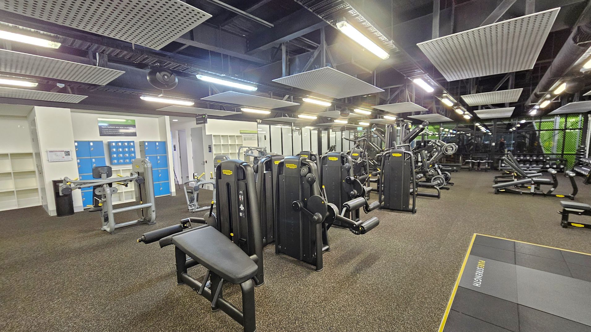 A spacious gym with various exercise machines. On the left, there are blue lockers and shelves against a light-coloured wall. In the centre, multiple black and grey workout machines are arranged in rows. To the right, more equipment, including benches and weights, is visible. The floor is covered with brown carpet tiles, and the ceiling features exposed beams and rectangular light fixtures. A spacious gym with various exercise machines. On the left, there are blue lockers and shelves against a light-coloured wall. In the centre, multiple black and grey workout machines are arranged in rows. To the right, more equipment, including benches and weights, is visible. The floor is covered with brown carpet tiles, and the ceiling features exposed beams and rectangular light fixtures.