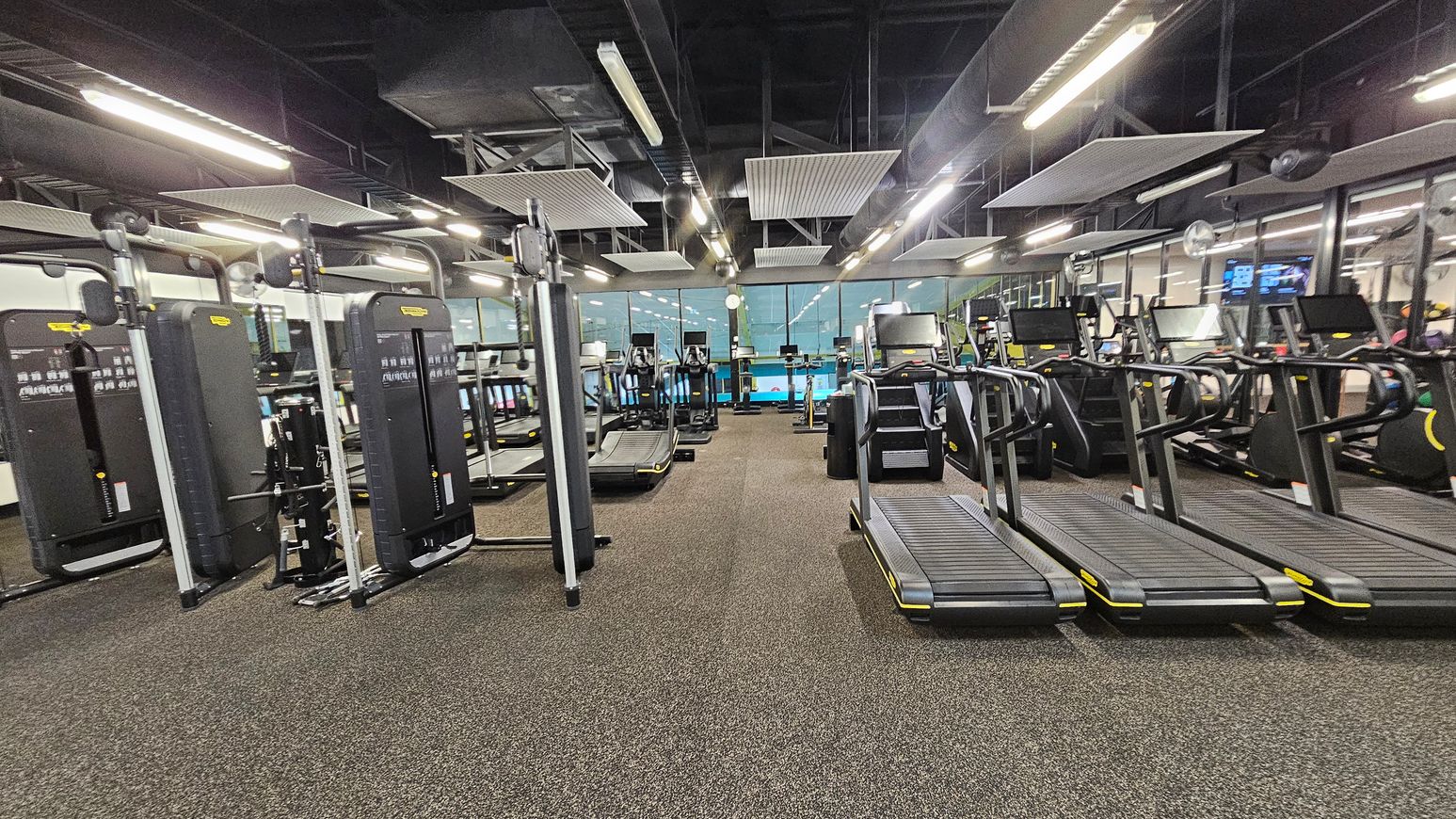 A gym with a variety of exercise equipment. On the left, there are several weight machines with black panels and metal frames. In the centre, there are multiple treadmills and elliptical machines lined up, facing large windows. On the right, more treadmills are visible. The floor is covered with a speckled brown and green carpet. The ceiling is high with exposed ductwork and rectangular light fixtures. A gym with a variety of exercise equipment. On the left, there are several weight machines with black panels and metal frames. In the centre, there are multiple treadmills and elliptical machines lined up, facing large windows. On the right, more treadmills are visible. The floor is covered with a speckled brown and green carpet. The ceiling is high with exposed ductwork and rectangular light fixtures.