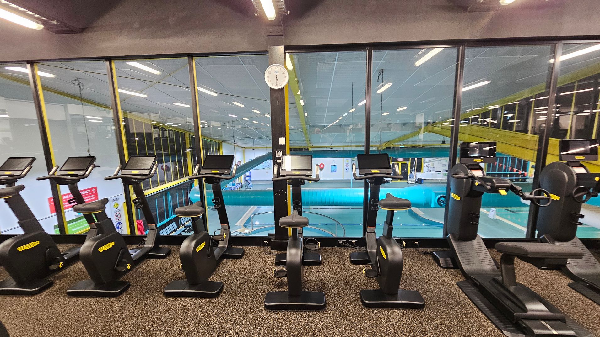 A row of stationary exercise bikes and recumbent bikes with digital screens is positioned on a speckled carpeted floor. Behind them, large glass windows reveal an indoor swimming pool area with a blue water slide and yellow railings. A white clock is mounted on a black pillar in the centre of the window. The ceiling has fluorescent lights, and the overall setting is a gym overlooking a pool area. A row of stationary exercise bikes and recumbent bikes with digital screens is positioned on a speckled carpeted floor. Behind them, large glass windows reveal an indoor swimming pool area with a blue water slide and yellow railings. A white clock is mounted on a black pillar in the centre of the window. The ceiling has fluorescent lights, and the overall setting is a gym overlooking a pool area.