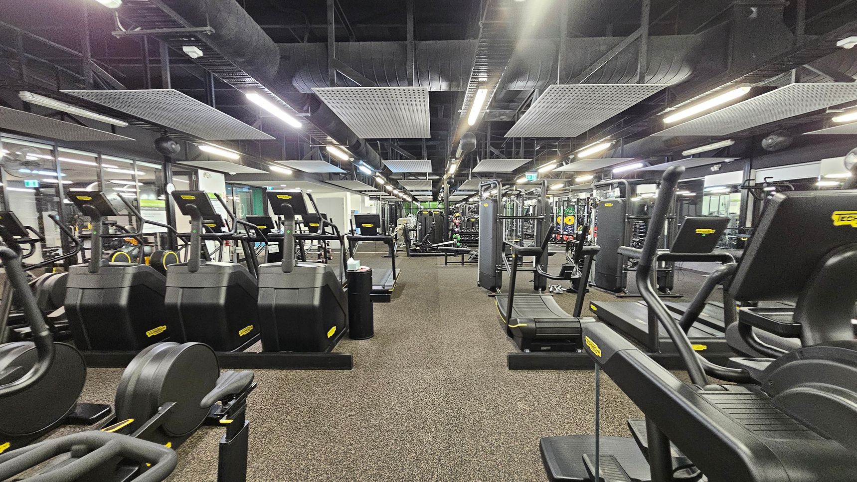 A spacious gym interior with rows of black exercise machines, including treadmills and ellipticals, on both sides. The floor is covered with brown carpet tiles. The ceiling is high with exposed ductwork and rectangular acoustic panels. Bright fluorescent lights illuminate the space. In the background, there are additional fitness equipment and weightlifting areas. Large mirrors line the walls, reflecting the equipment and enhancing the sense of space. A spacious gym interior with rows of black exercise machines, including treadmills and ellipticals, on both sides. The floor is covered with brown carpet tiles. The ceiling is high with exposed ductwork and rectangular acoustic panels. Bright fluorescent lights illuminate the space. In the background, there are additional fitness equipment and weightlifting areas. Large mirrors line the walls, reflecting the equipment and enhancing the sense of space.