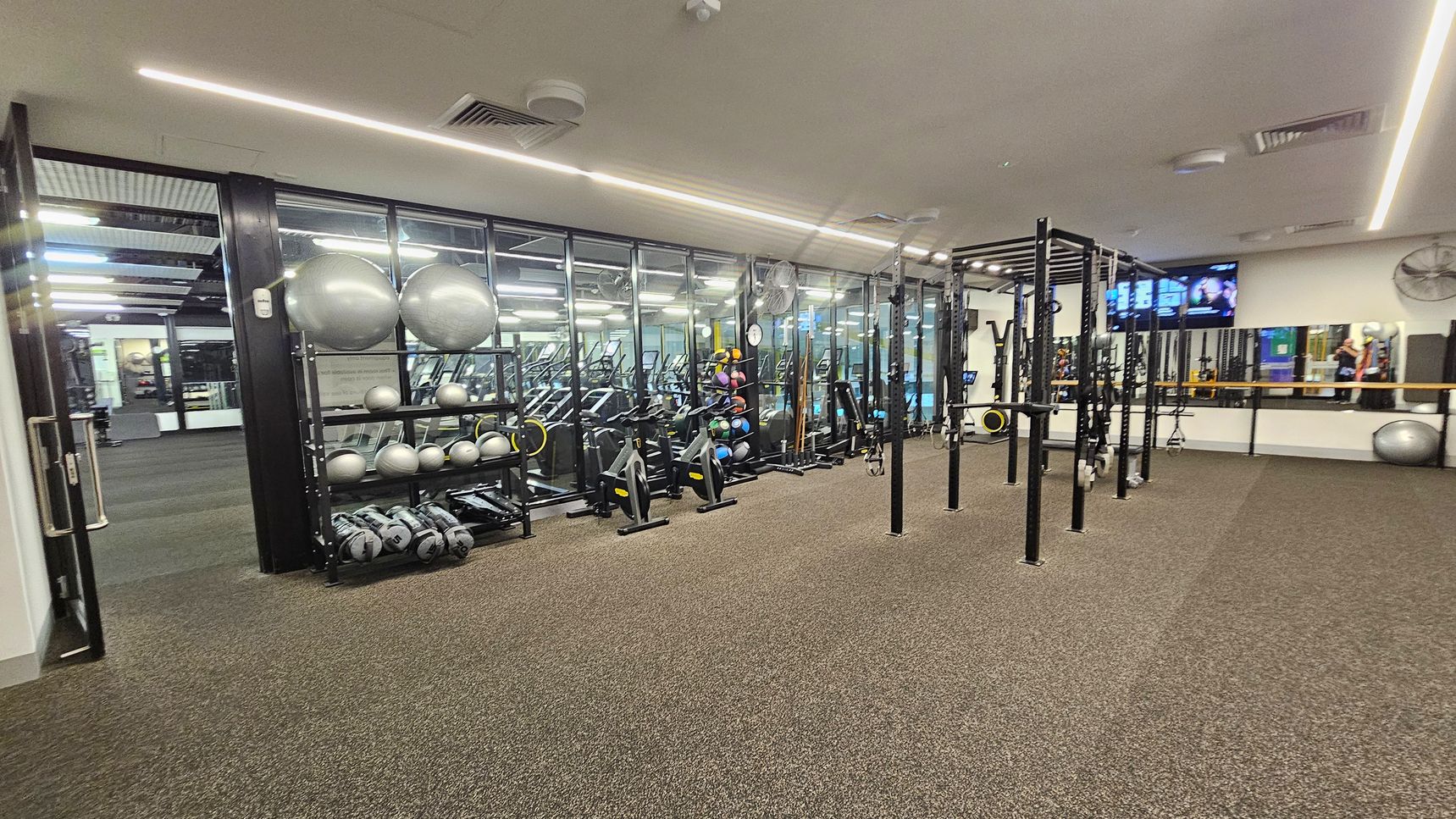 A gym with a brown carpeted floor and a ceiling with recessed lighting. On the left, there is a rack with silver exercise balls and medicine balls. In the middle, there are stationary bikes and a large metal frame for exercises. On the right, a wall-mounted TV and a mirror reflect the gym equipment. The background features glass walls separating this area from another section of the gym. A gym with a brown carpeted floor and a ceiling with recessed lighting. On the left, there is a rack with silver exercise balls and medicine balls. In the middle, there are stationary bikes and a large metal frame for exercises. On the right, a wall-mounted TV and a mirror reflect the gym equipment. The background features glass walls separating this area from another section of the gym.