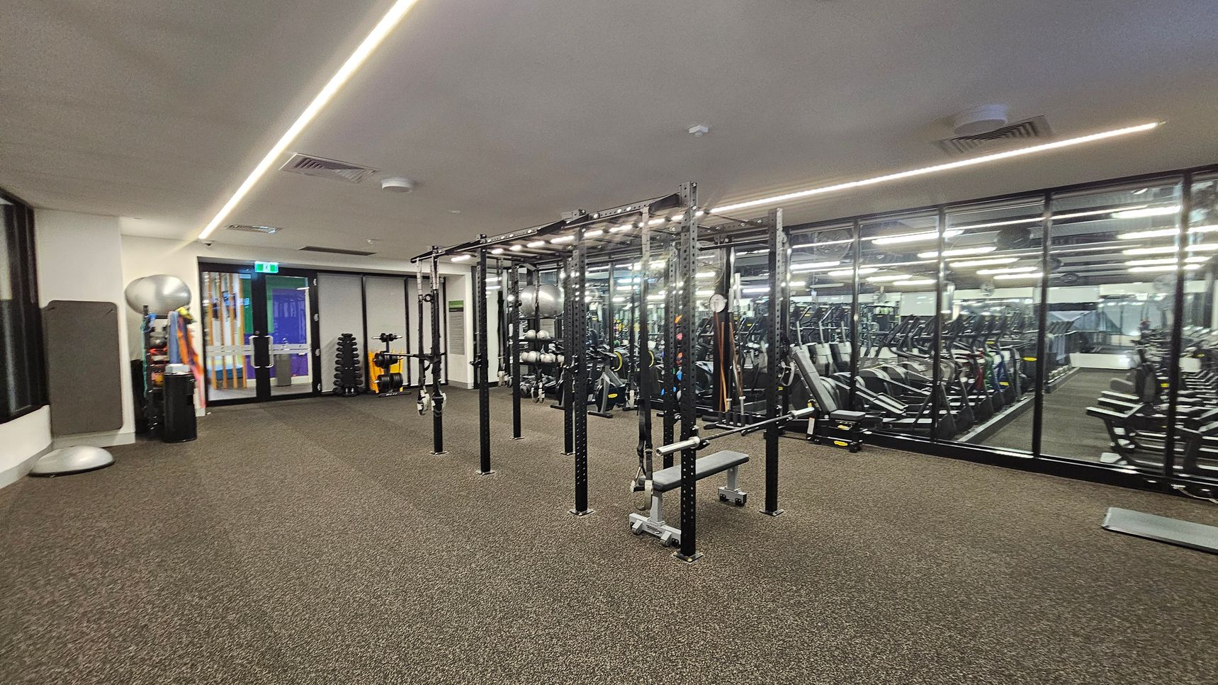 A gym with a brown carpeted floor and a white ceiling featuring linear lights. On the left, there are exercise balls, mats, and a doorway. In the centre, a large metal frame structure with various gym equipment like benches and straps. On the right, a mirrored wall reflects rows of treadmills and other cardio machines. A gym with a brown carpeted floor and a white ceiling featuring linear lights. On the left, there are exercise balls, mats, and a doorway. In the centre, a large metal frame structure with various gym equipment like benches and straps. On the right, a mirrored wall reflects rows of treadmills and other cardio machines.
