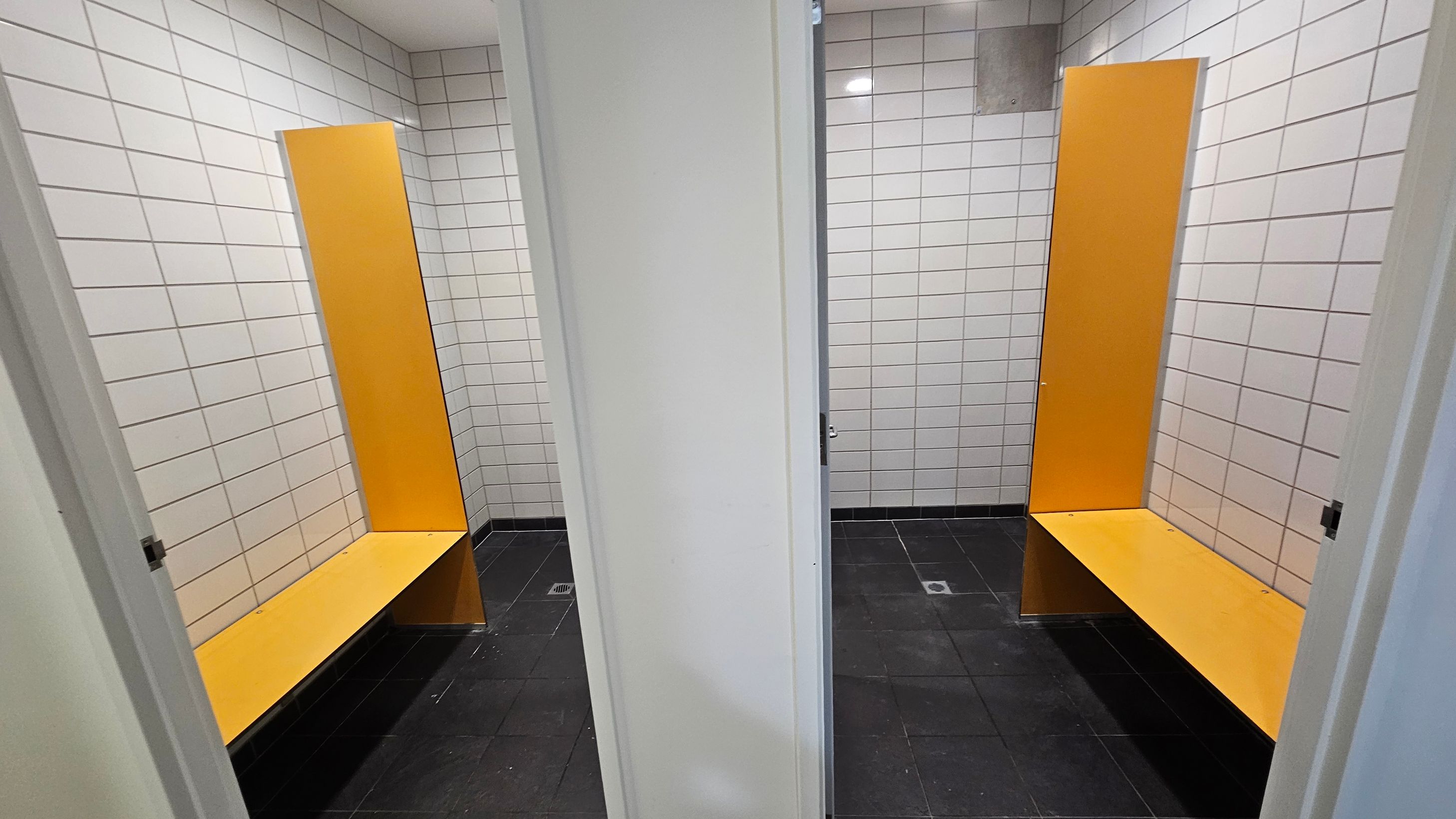 Two changing cubicles are visible, separated by a white partition. Each cubicle has white tiled walls and a black tiled floor. Inside each cubicle, there is a bright orange bench with a matching vertical panel behind it. The ceiling is plain white with recessed lighting. Two changing cubicles are visible, separated by a white partition. Each cubicle has white tiled walls and a black tiled floor. Inside each cubicle, there is a bright orange bench with a matching vertical panel behind it. The ceiling is plain white with recessed lighting.