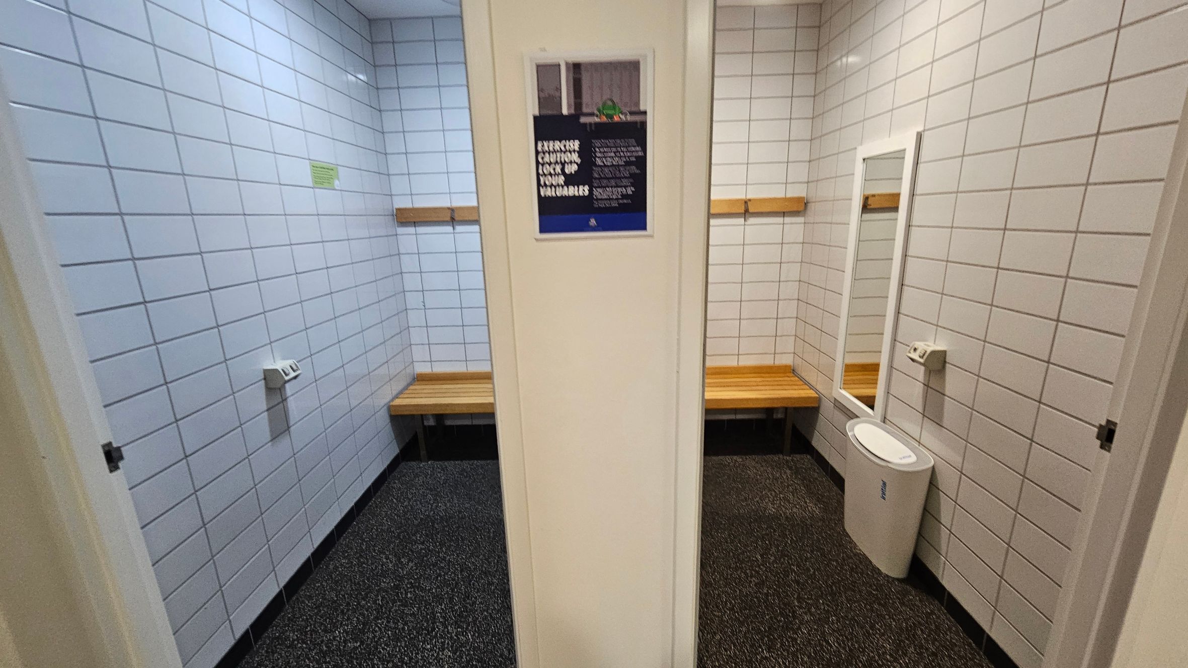 A changing room with white tiled walls and a speckled floor. On the left, there is a wooden bench beneath a coat hook. In the centre, a wall with a sign. On the right, another wooden bench, a full-length mirror, and a white bin. The ceiling is plain with recessed lighting. A changing room with white tiled walls and a speckled floor. On the left, there is a wooden bench beneath a coat hook. In the centre, a wall with a sign. On the right, another wooden bench, a full-length mirror, and a white bin. The ceiling is plain with recessed lighting.