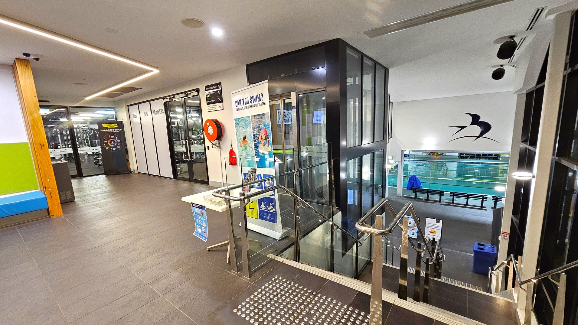 A modern indoor facility with a tiled floor. On the left, there's a gym area visible through glass doors, with a sign about trainers. A red fire hose reel is mounted on the wall. In the centre, a lift with glass panels is adjacent to a staircase with metal railings leading down. A table with informational posters is nearby. On the right, a swimming pool area is visible below, with a large bird logo on the wall. The ceiling has recessed lighting and a sleek design. A modern indoor facility with a tiled floor. On the left, there's a gym area visible through glass doors, with a sign about trainers. A red fire hose reel is mounted on the wall. In the centre, a lift with glass panels is adjacent to a staircase with metal railings leading down. A table with informational posters is nearby. On the right, a swimming pool area is visible below, with a large bird logo on the wall. The ceiling has recessed lighting and a sleek design.