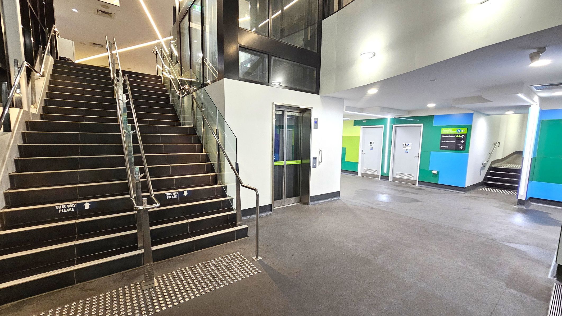 A modern indoor space with a staircase on the left featuring metal railings and glass panels. The stairs have signs reading "This Way Please." To the right, there is a lift with a green stripe on the doors. The walls are painted in shades of blue and green, with doors leading to other rooms. A sign indicates directions to changing rooms and other facilities. The floor is grey, and the ceiling has recessed lighting. A modern indoor space with a staircase on the left featuring metal railings and glass panels. The stairs have signs reading "This Way Please." To the right, there is a lift with a green stripe on the doors. The walls are painted in shades of blue and green, with doors leading to other rooms. A sign indicates directions to changing rooms and other facilities. The floor is grey, and the ceiling has recessed lighting.