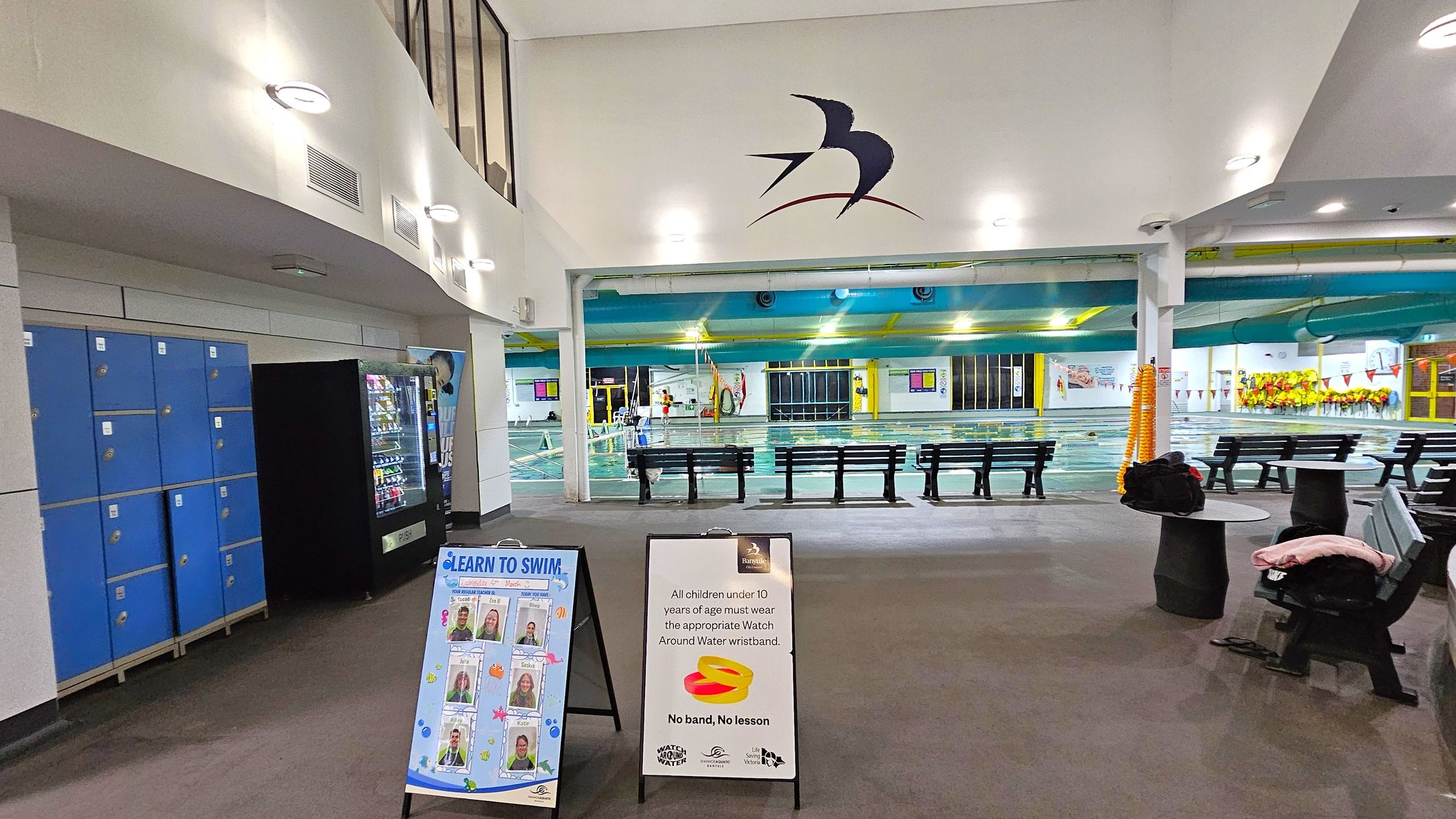 Indoor swimming pool area with blue lockers and a vending machine on the left. Two signs are in the foreground, one promoting swimming lessons and the other about wristband requirements for children. In the centre, benches face the pool, which is visible in the background. The ceiling is white with circular lights, and a bird logo is on the wall above the pool entrance. The floor is carpeted, and there are tables with clothes on the right. Indoor swimming pool area with blue lockers and a vending machine on the left. Two signs are in the foreground, one promoting swimming lessons and the other about wristband requirements for children. In the centre, benches face the pool, which is visible in the background. The ceiling is white with circular lights, and a bird logo is on the wall above the pool entrance. The floor is carpeted, and there are tables with clothes on the right.