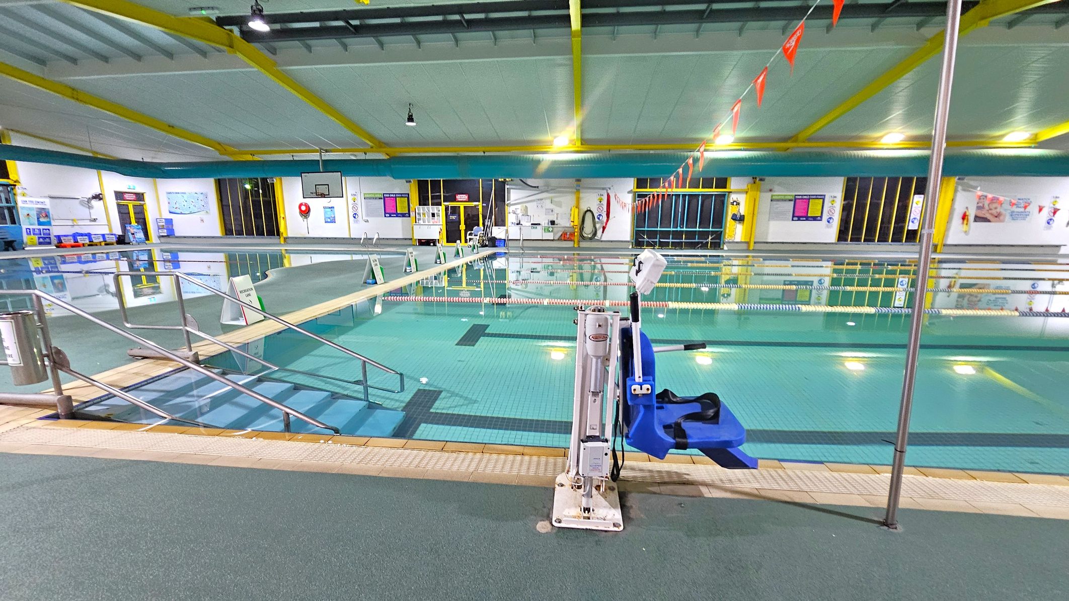 An indoor swimming pool with clear water and lane dividers. In the foreground, a blue pool lift is attached to the poolside. A ramp with metal railings leads into the water on the left. The ceiling has yellow beams and is well-lit with overhead lights. The walls are adorned with various signs and posters. A basketball hoop is visible in the background, and there are emergency equipment and doors along the walls. The floor around the pool is covered with non-slip material. An indoor swimming pool with clear water and lane dividers. In the foreground, a blue pool lift is attached to the poolside. A ramp with metal railings leads into the water on the left. The ceiling has yellow beams and is well-lit with overhead lights. The walls are adorned with various signs and posters. A basketball hoop is visible in the background, and there are emergency equipment and doors along the walls. The floor around the pool is covered with non-slip material.