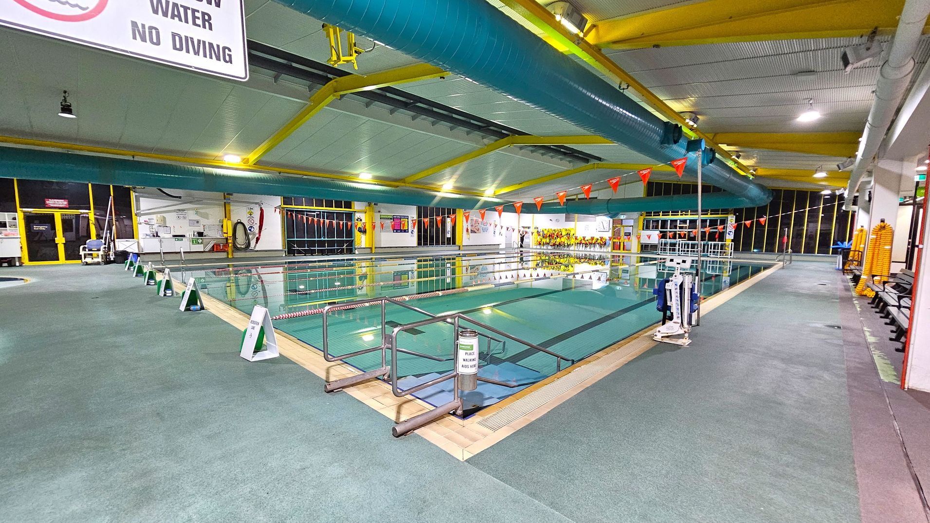 An indoor swimming pool with lanes marked by ropes and orange triangular flags overhead. The pool is surrounded by a green non-slip floor. On the left, there are signs warning of shallow water and no diving. The ceiling features yellow beams and large blue ventilation ducts. On the right, there are benches and stacked yellow flotation devices. The far end of the pool area has large windows and various safety equipment mounted on the walls. An indoor swimming pool with lanes marked by ropes and orange triangular flags overhead. The pool is surrounded by a green non-slip floor. On the left, there are signs warning of shallow water and no diving. The ceiling features yellow beams and large blue ventilation ducts. On the right, there are benches and stacked yellow flotation devices. The far end of the pool area has large windows and various safety equipment mounted on the walls.