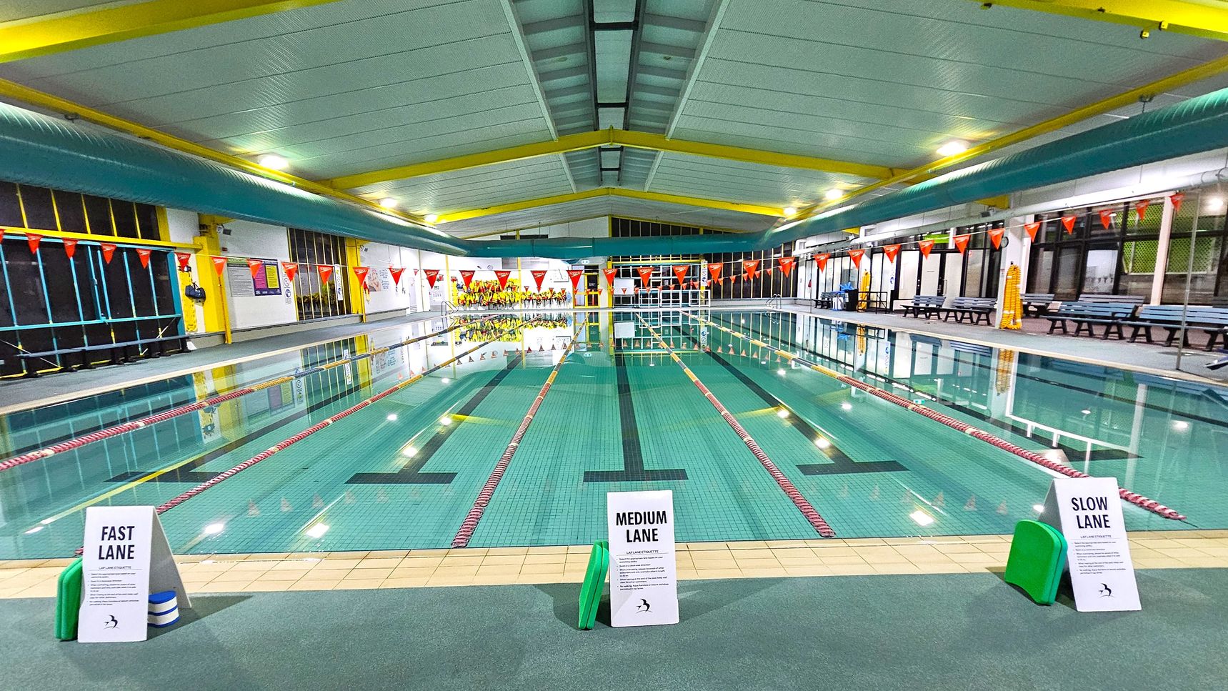An indoor swimming pool with three lanes marked by signs: "Fast Lane" on the left, "Medium Lane" in the centre, and "Slow Lane" on the right. The pool is bordered by red and white lane dividers. The ceiling is white with yellow beams, and the walls have large windows and benches along the sides. Orange triangular flags hang above the pool. The floor around the pool is tiled, and there are green kickboards near each lane sign. An indoor swimming pool with three lanes marked by signs: "Fast Lane" on the left, "Medium Lane" in the centre, and "Slow Lane" on the right. The pool is bordered by red and white lane dividers. The ceiling is white with yellow beams, and the walls have large windows and benches along the sides. Orange triangular flags hang above the pool. The floor around the pool is tiled, and there are green kickboards near each lane sign.