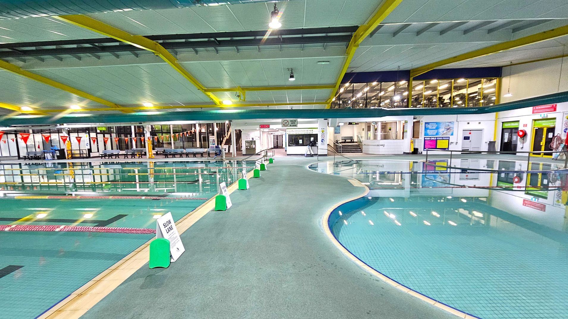 Indoor swimming pool area with a wide walkway in the centre, bordered by pools on both sides. The left side features a lap pool with lane dividers and triangular flags above. The right side has a smaller, shallow pool. Green and white signs are placed along the walkway. The background shows a glass-walled gym area and various doors and signs. The ceiling is white with yellow beams and overhead lights. Indoor swimming pool area with a wide walkway in the centre, bordered by pools on both sides. The left side features a lap pool with lane dividers and triangular flags above. The right side has a smaller, shallow pool. Green and white signs are placed along the walkway. The background shows a glass-walled gym area and various doors and signs. The ceiling is white with yellow beams and overhead lights.