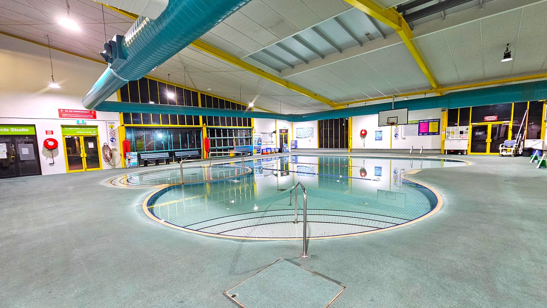 Indoor swimming pool area with a circular pool in the foreground, surrounded by a grey tiled floor. A metal handrail leads into the pool. Large windows with yellow frames line the walls, and a blue ventilation duct runs across the ceiling. On the left, there are doors labelled "Cycle Studio" and emergency equipment like a fire hose. On the right, a basketball hoop is mounted on the wall. Various signs and safety equipment are visible around the pool area. Indoor swimming pool area with a circular pool in the foreground, surrounded by a grey tiled floor. A metal handrail leads into the pool. Large windows with yellow frames line the walls, and a blue ventilation duct runs across the ceiling. On the left, there are doors labelled "Cycle Studio" and emergency equipment like a fire hose. On the right, a basketball hoop is mounted on the wall. Various signs and safety equipment are visible around the pool area.