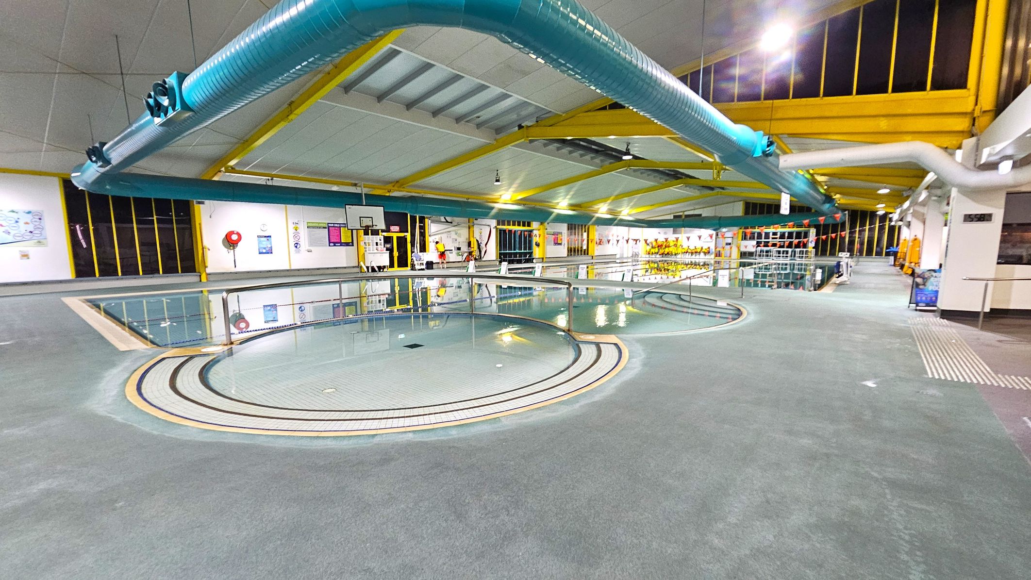Indoor swimming pool area with a circular shallow pool in the foreground. The floor is grey and textured. In the background, a larger pool is visible with lane dividers and a basketball hoop. The ceiling is supported by yellow beams, and large blue ventilation ducts run across it. Walls have windows with yellow frames and various posters. On the right, there are shelves with equipment and a walkway leading further into the facility. Indoor swimming pool area with a circular shallow pool in the foreground. The floor is grey and textured. In the background, a larger pool is visible with lane dividers and a basketball hoop. The ceiling is supported by yellow beams, and large blue ventilation ducts run across it. Walls have windows with yellow frames and various posters. On the right, there are shelves with equipment and a walkway leading further into the facility.