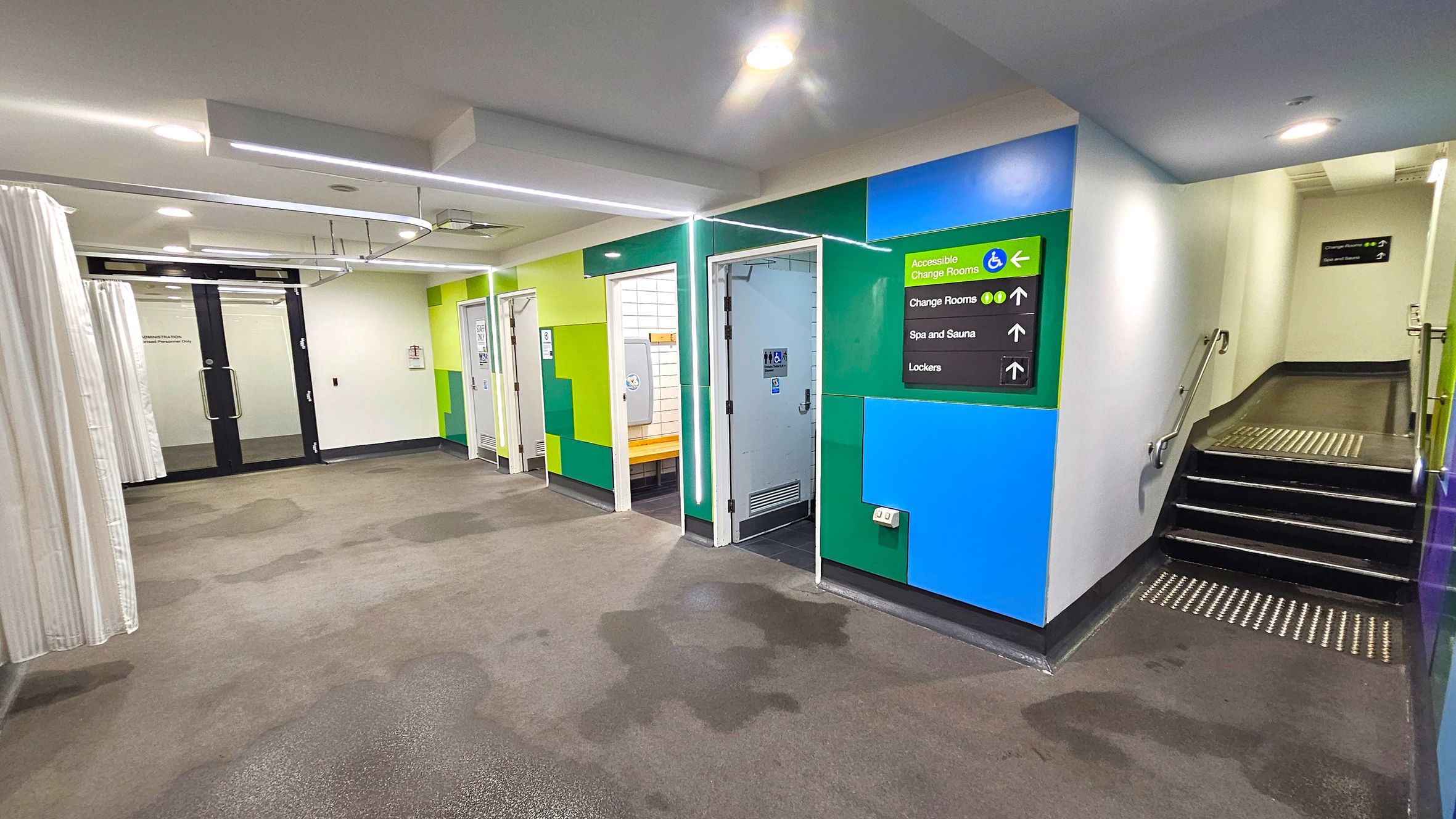 A corridor with a carpeted floor and a white ceiling featuring recessed lighting. On the left, there are glass doors with curtains. In the centre, there are several doors leading to changing rooms, with colourful green and blue walls. A sign indicates directions to accessible change rooms, spa and sauna, and lockers. On the right, a staircase with a handrail leads upwards. A corridor with a carpeted floor and a white ceiling featuring recessed lighting. On the left, there are glass doors with curtains. In the centre, there are several doors leading to changing rooms, with colourful green and blue walls. A sign indicates directions to accessible change rooms, spa and sauna, and lockers. On the right, a staircase with a handrail leads upwards.