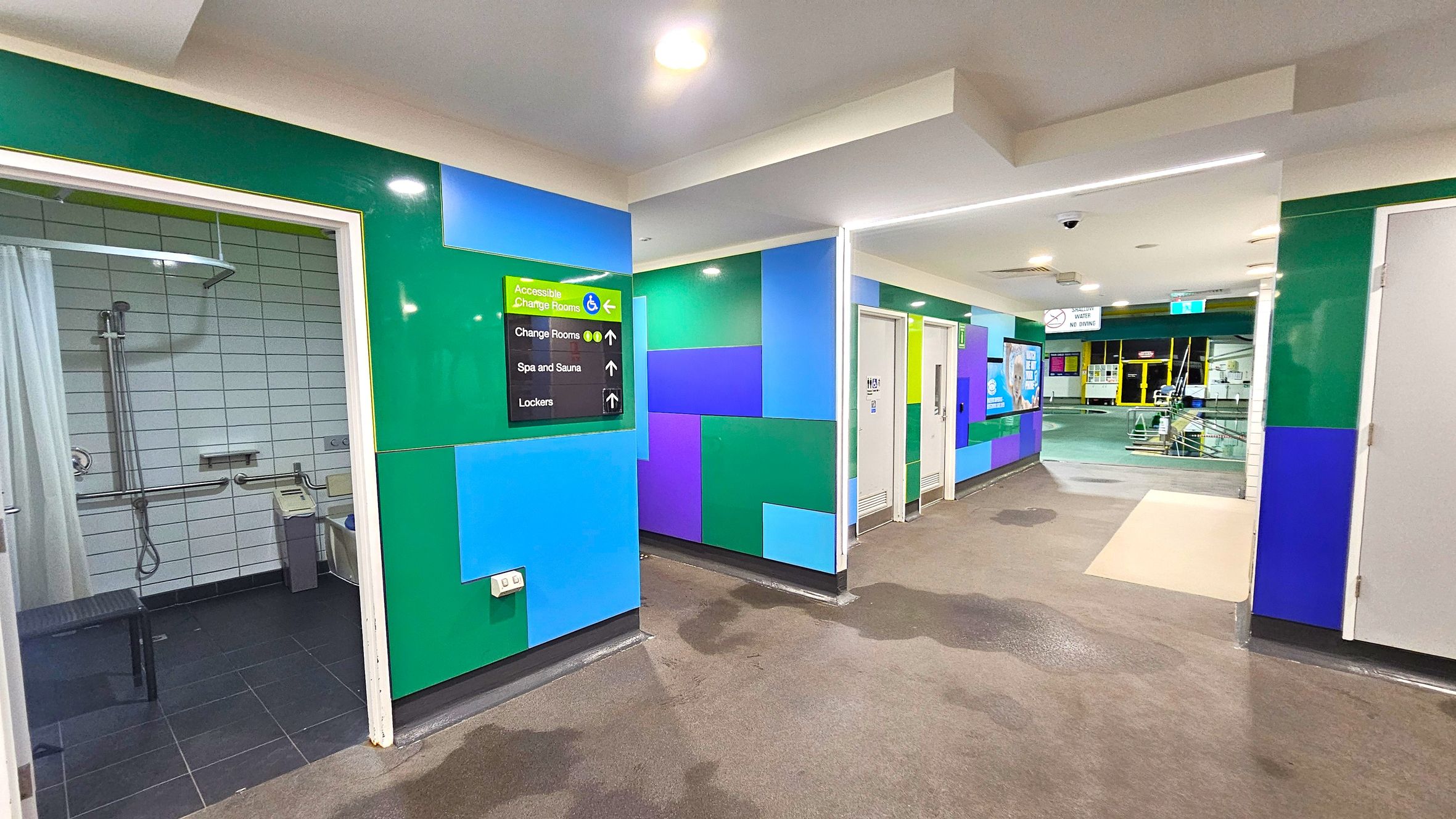 On the left, there is an accessible shower room with a curtain, grab bars, and a showerhead. The walls are tiled in white. In the centre, a colourful wall with green, blue, and purple panels features a sign indicating directions to accessible change rooms, spa and sauna, and lockers. On the right, there are doors leading to other facilities. The floor is dark with some wet patches, and the ceiling is white with recessed lighting. In the background, there is a view of a pool area. On the left, there is an accessible shower room with a curtain, grab bars, and a showerhead. The walls are tiled in white. In the centre, a colourful wall with green, blue, and purple panels features a sign indicating directions to accessible change rooms, spa and sauna, and lockers. On the right, there are doors leading to other facilities. The floor is dark with some wet patches, and the ceiling is white with recessed lighting. In the background, there is a view of a pool area.