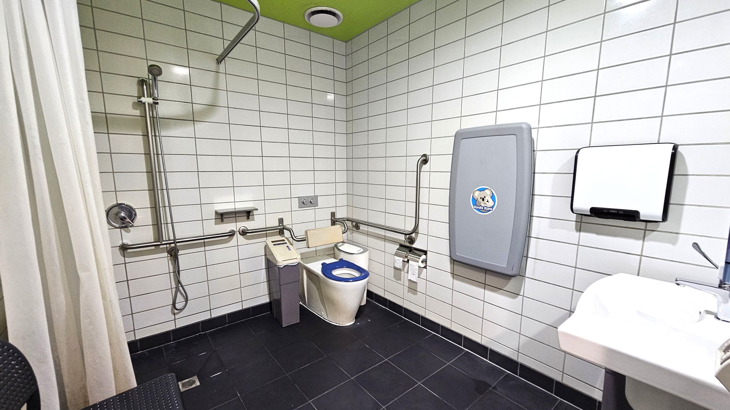 A public accessible bathroom with white tiled walls and a green ceiling. On the left, there is a shower area with a curtain and a handheld showerhead attached to a vertical rail. In the centre, a toilet with a blue seat is equipped with support rails and a small control panel. On the right, a baby changing station is mounted on the wall above a paper towel dispenser. A white sink with a lever tap is visible in the foreground. The floor is covered with dark tiles. A public accessible bathroom with white tiled walls and a green ceiling. On the left, there is a shower area with a curtain and a handheld showerhead attached to a vertical rail. In the centre, a toilet with a blue seat is equipped with support rails and a small control panel. On the right, a baby changing station is mounted on the wall above a paper towel dispenser. A white sink with a lever tap is visible in the foreground. The floor is covered with dark tiles.